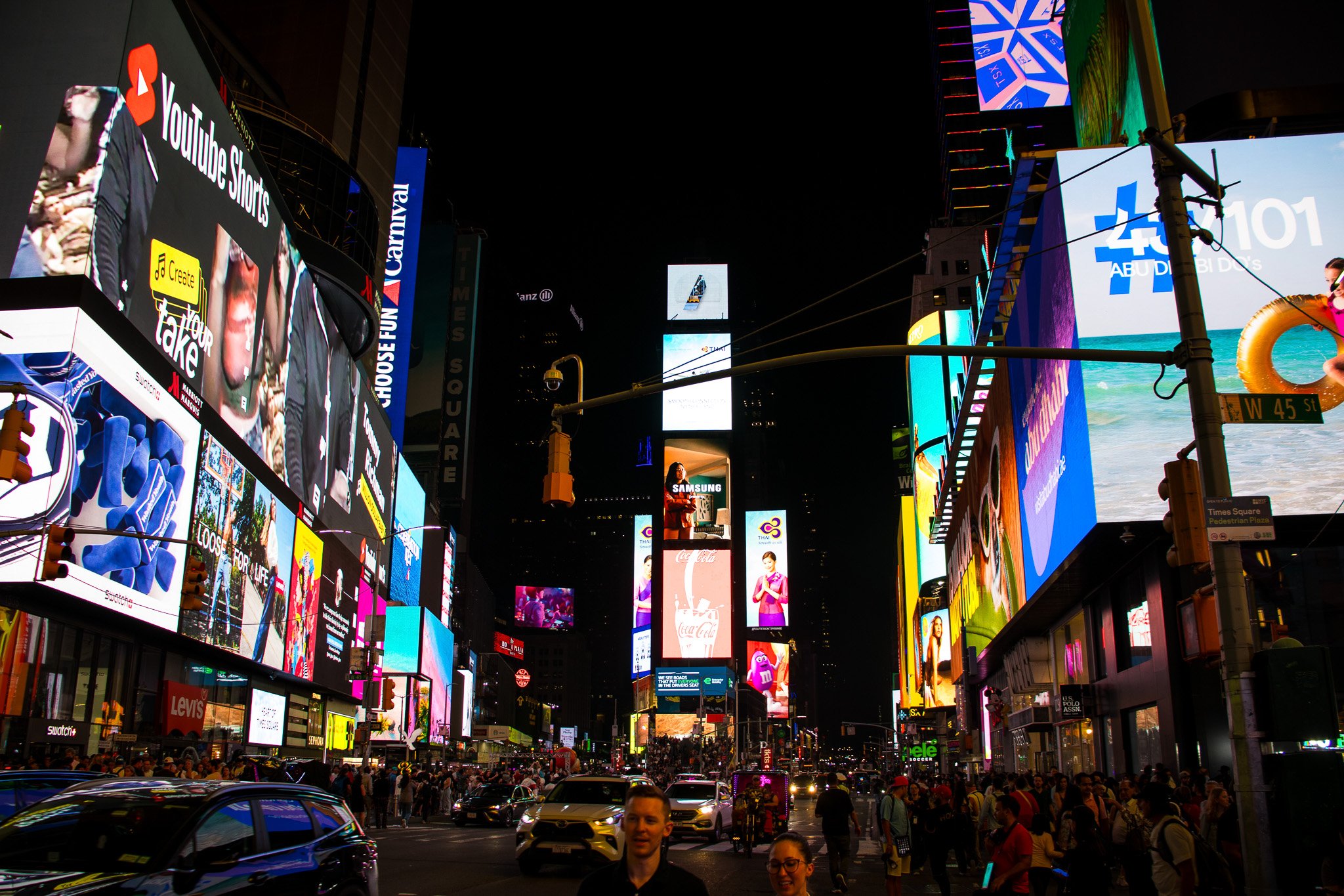 The image depicts Times Square in New York City at night, filled with colorful illuminated billboards and electronic advertisements. There is a crowd of people walking on the streets, and cars are visible in the foreground. Skyscrapers with various a
