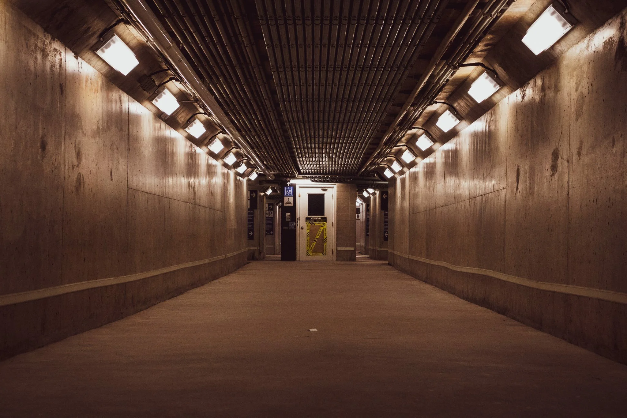 Underground tunnel or corridor with beige walls and ceiling pipes, illuminated by overhead fluorescent lights, leading to a door at the end with caution tape and warning signs.