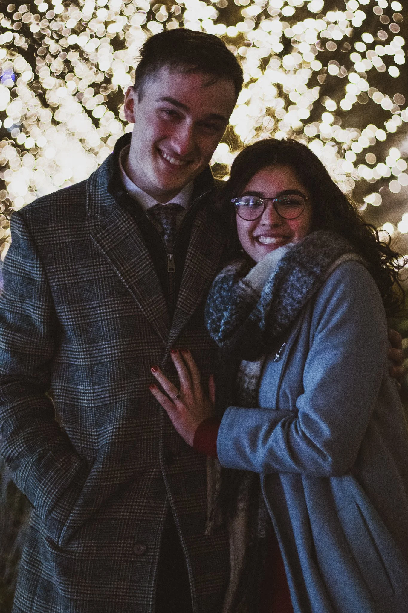 Two young people, a man and a woman, smiling and standing close together in front of a background of blurred lights, likely at night, with the woman showing a hand with a ring.