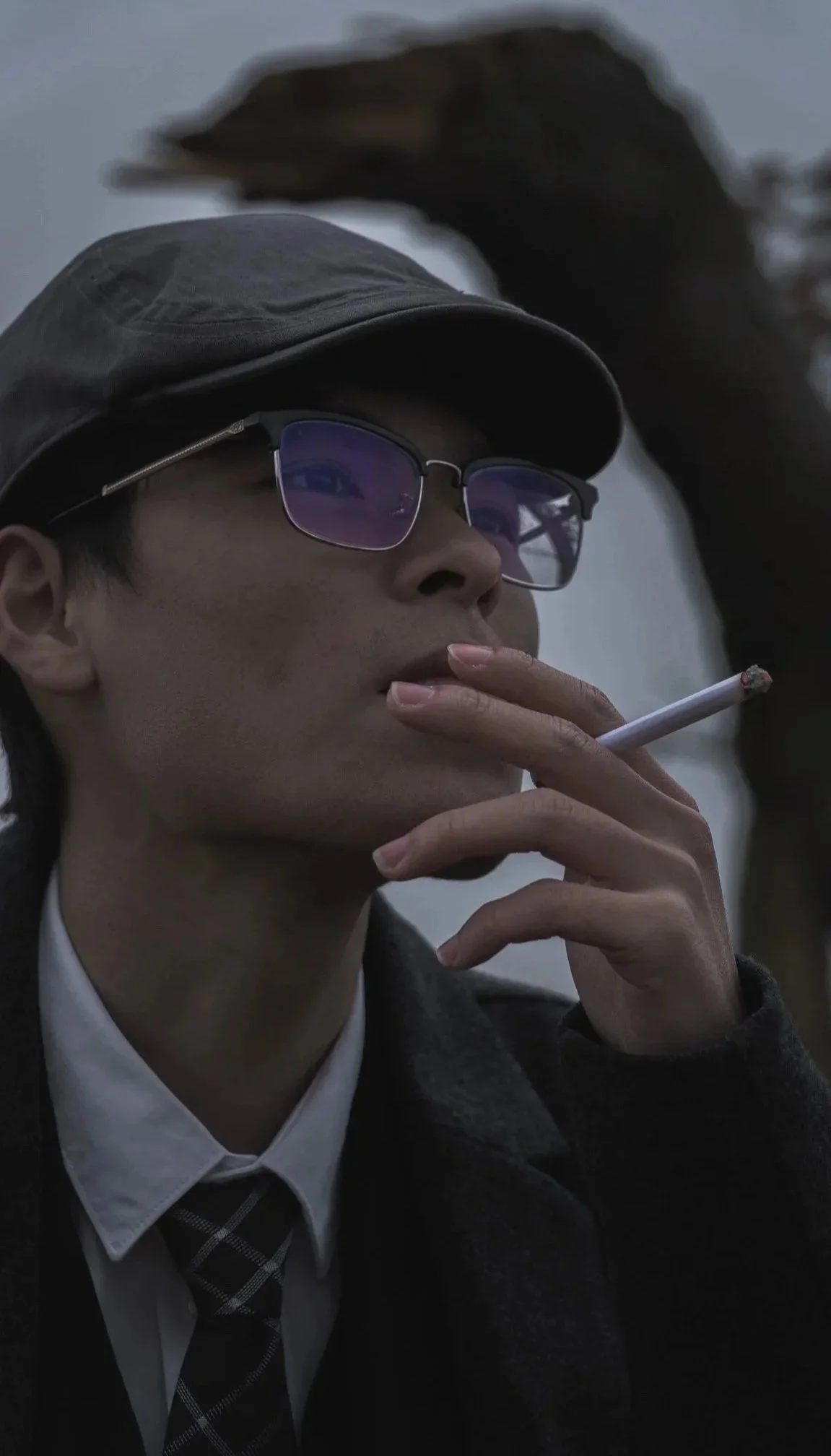 A person wearing glasses, a cap, a coat, a white shirt, and a tie, holding a cigarette near their lips.