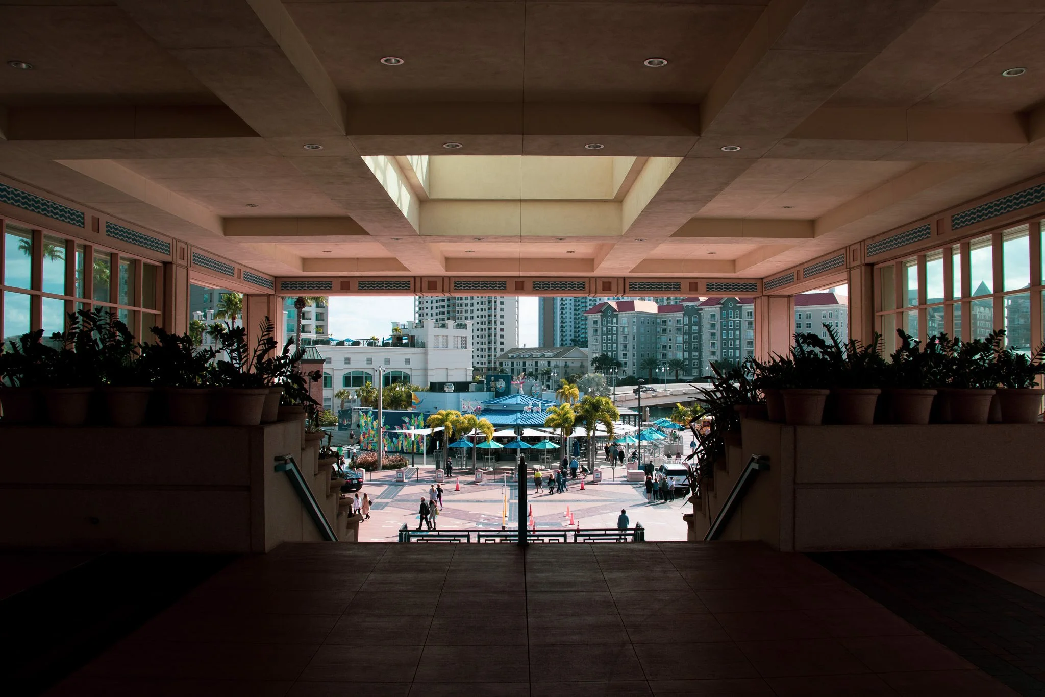 View of a busy outdoor area from inside a building, framed by a large open archway with a ceiling featuring a skylight. People are walking around, and there are high-rise buildings in the background.