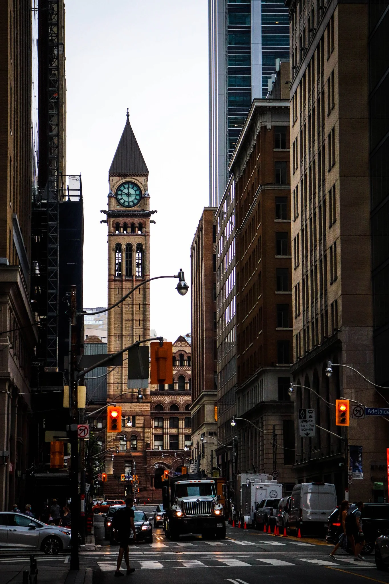 City street scene with tall buildings and a historic clock tower in the background. Traffic lights and vehicles, including a large truck, are visible at the intersection, with pedestrians walking on the crosswalks.