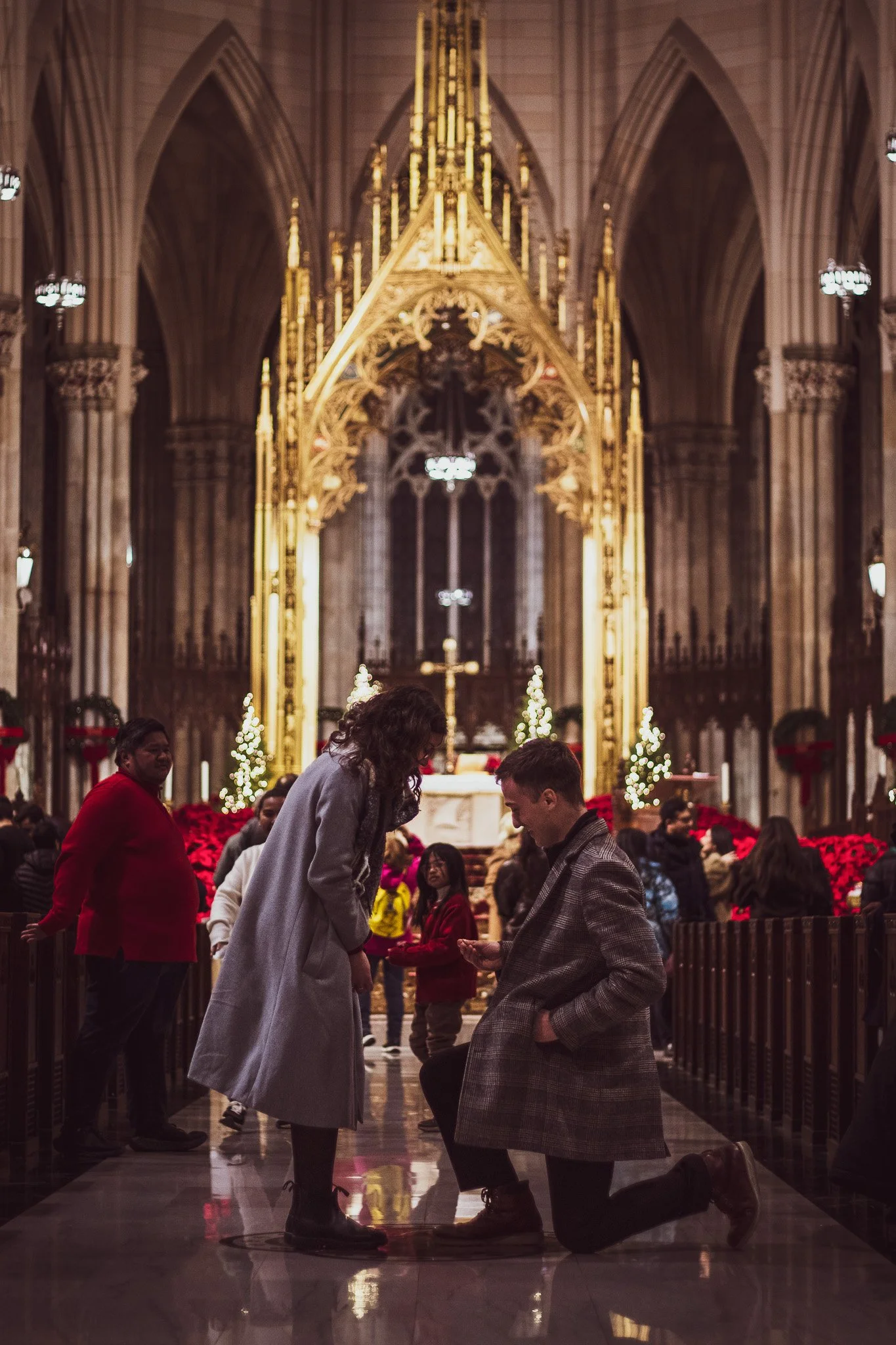 People in a church, with two individuals kneeling and holding hands in front of others, surrounded by festive Christmas decorations and lights.