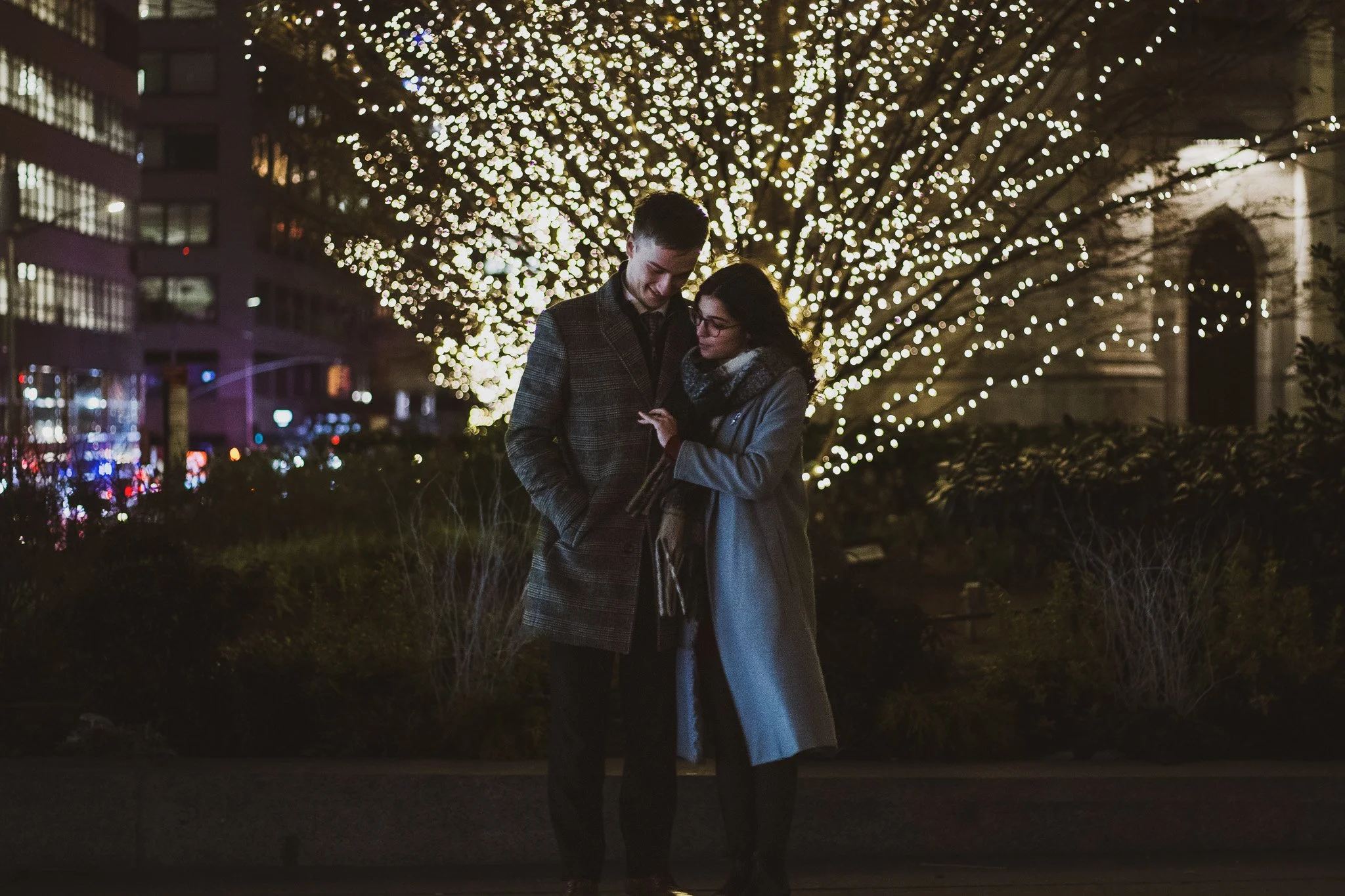 A couple standing close together and smiling at each other outdoors at night, with a large illuminated Christmas tree in the background.
