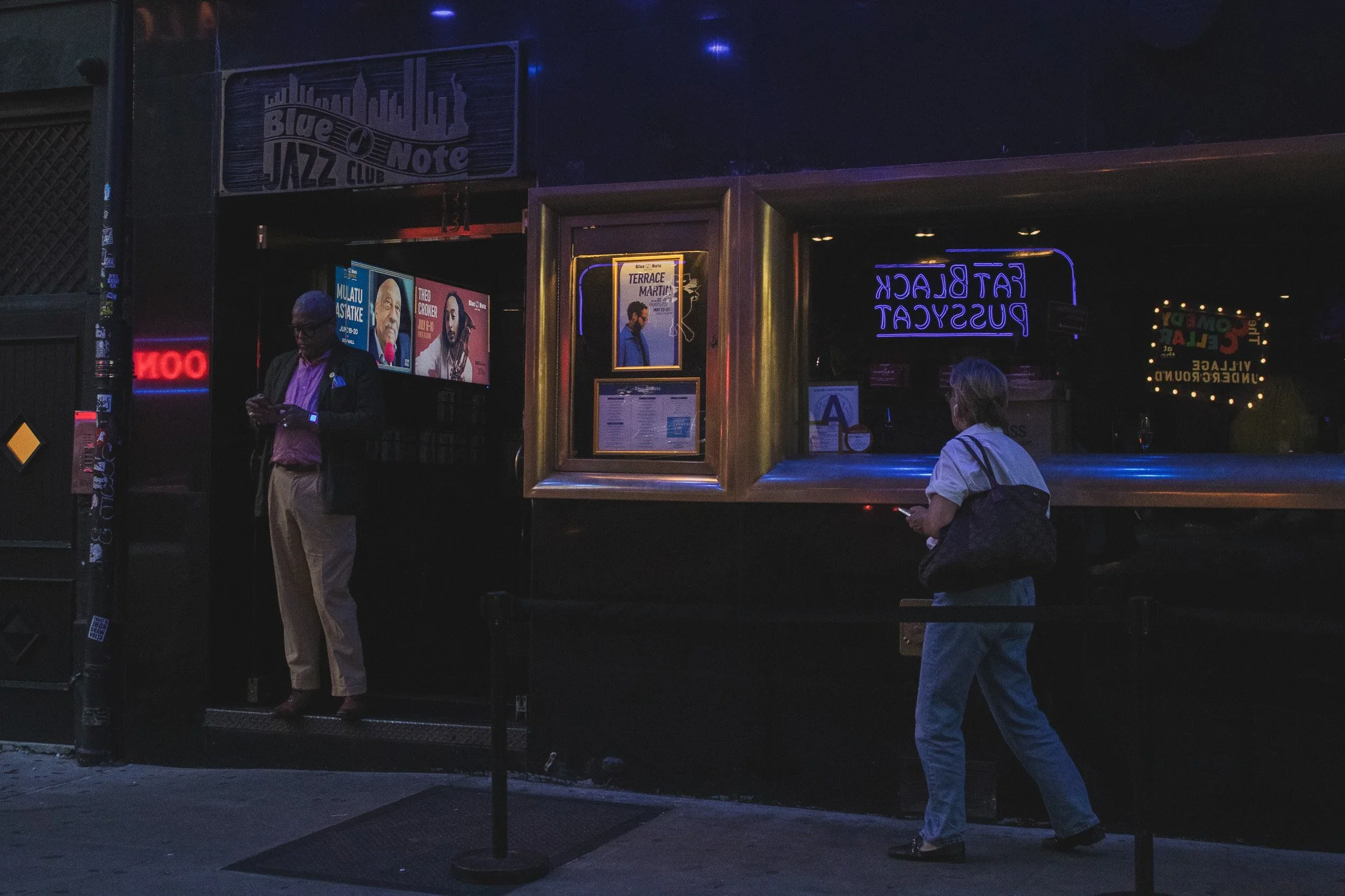 Nighttime scene outside a jazz club with two people standing near the entrance. A neon sign reads 'Fat Black Pussycat.' There are posters on the wall advertising upcoming performances. One person appears to be using a phone, and the other is standing