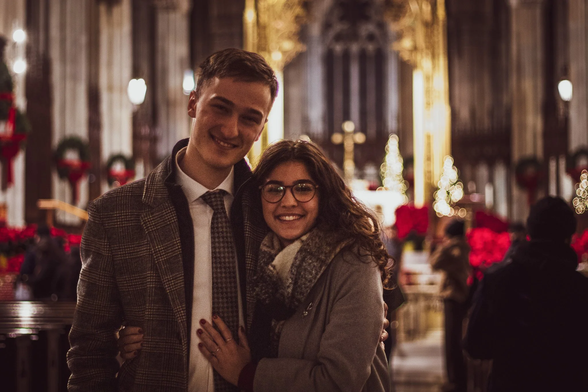 Two smiling people, a man and a woman, standing close together in what appears to be a decorated church or cathedral, with Christmas-themed decorations and warm lighting in the background.