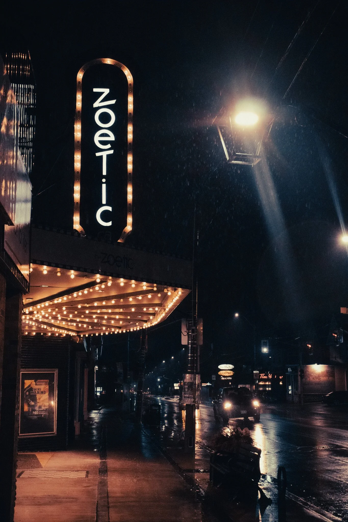 Nighttime street scene with a theater marquee displaying the word 'Zodiac' in lights, wet pavement reflecting streetlights, and a car with headlights on.