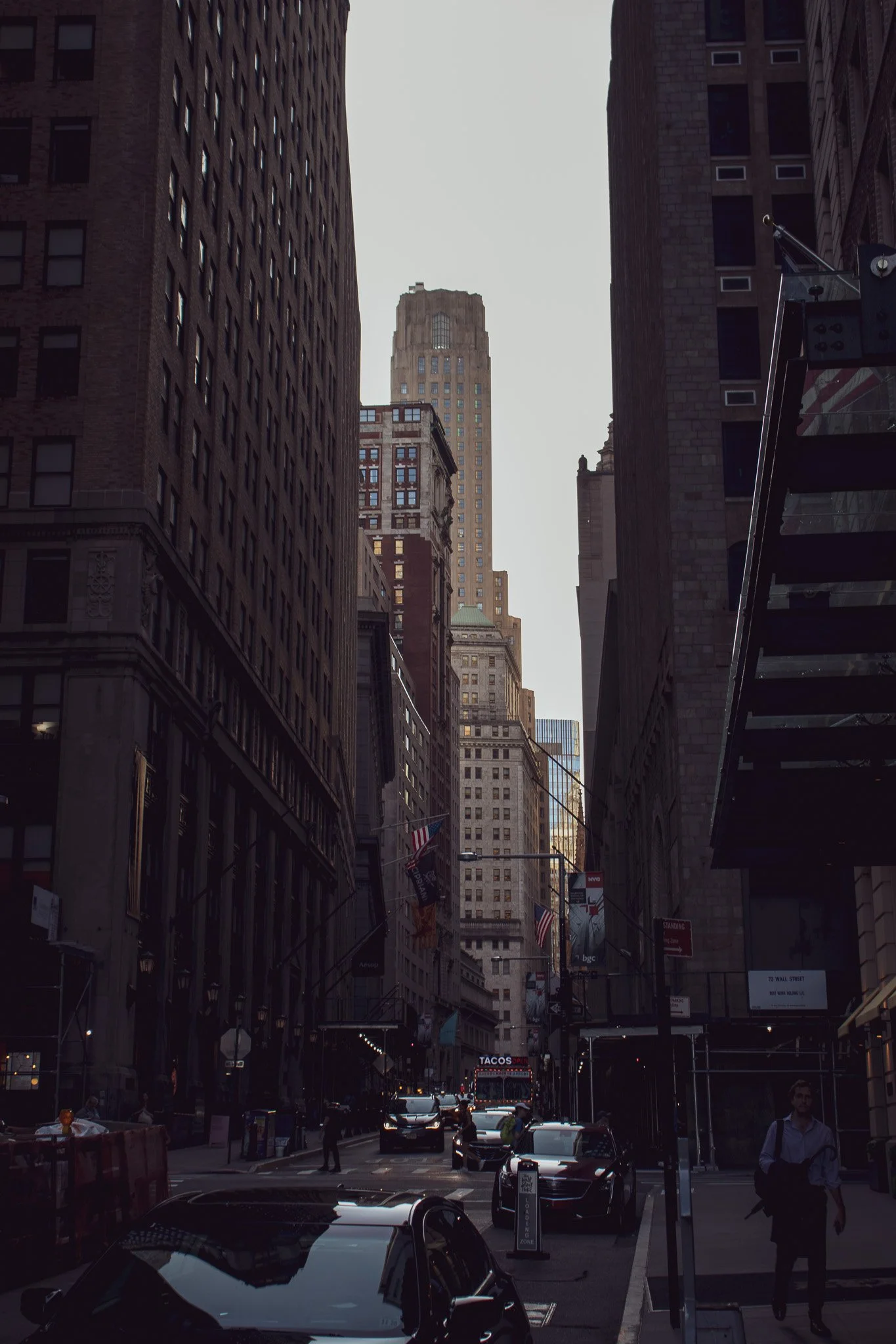 City street scene with tall buildings, cars parked, and pedestrians walking. A sign for tacos and flags are visible. The sky is overcast.