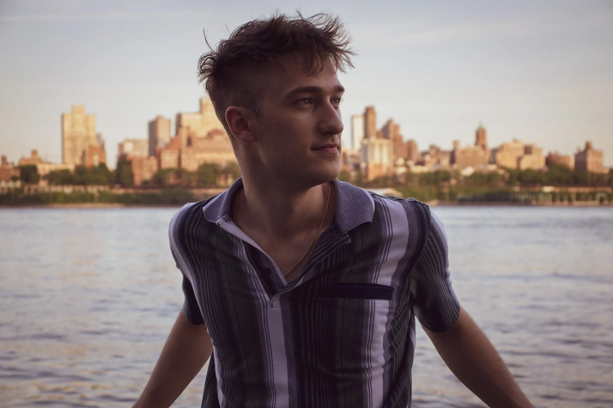A young man with tousled brown hair looking to the side by a body of water with a city skyline in the background during sunset.