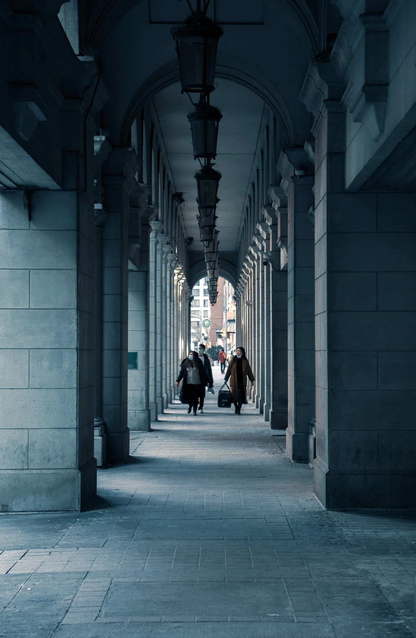 A covered walkway with stone columns and arches, lined with hanging lantern-style lights, with four people walking underneath. Two women in the center are wearing masks, one carrying a suitcase, the other holding a bag.