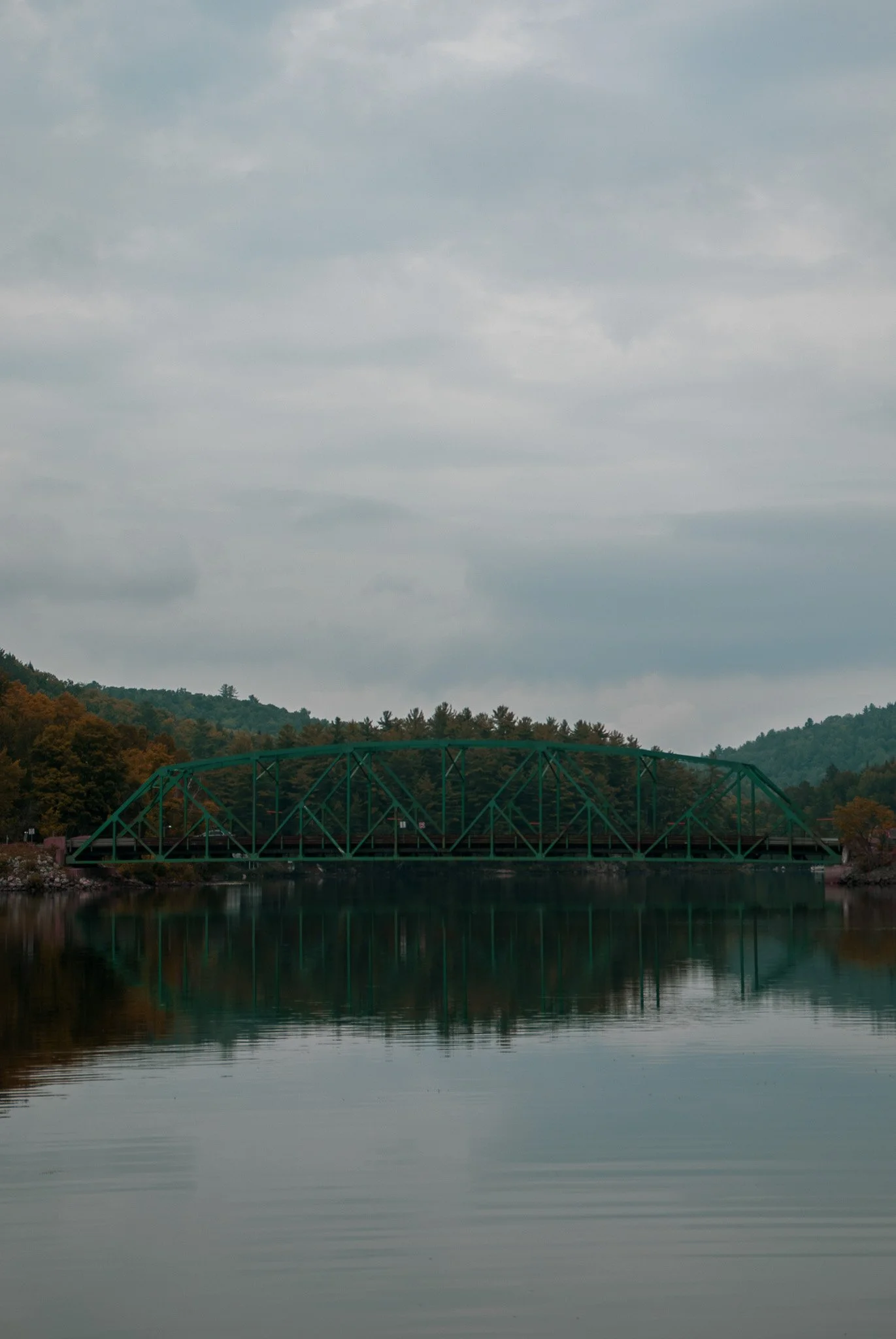 Green metal bridge over a calm body of water with forested hills and a cloudy sky in the background.