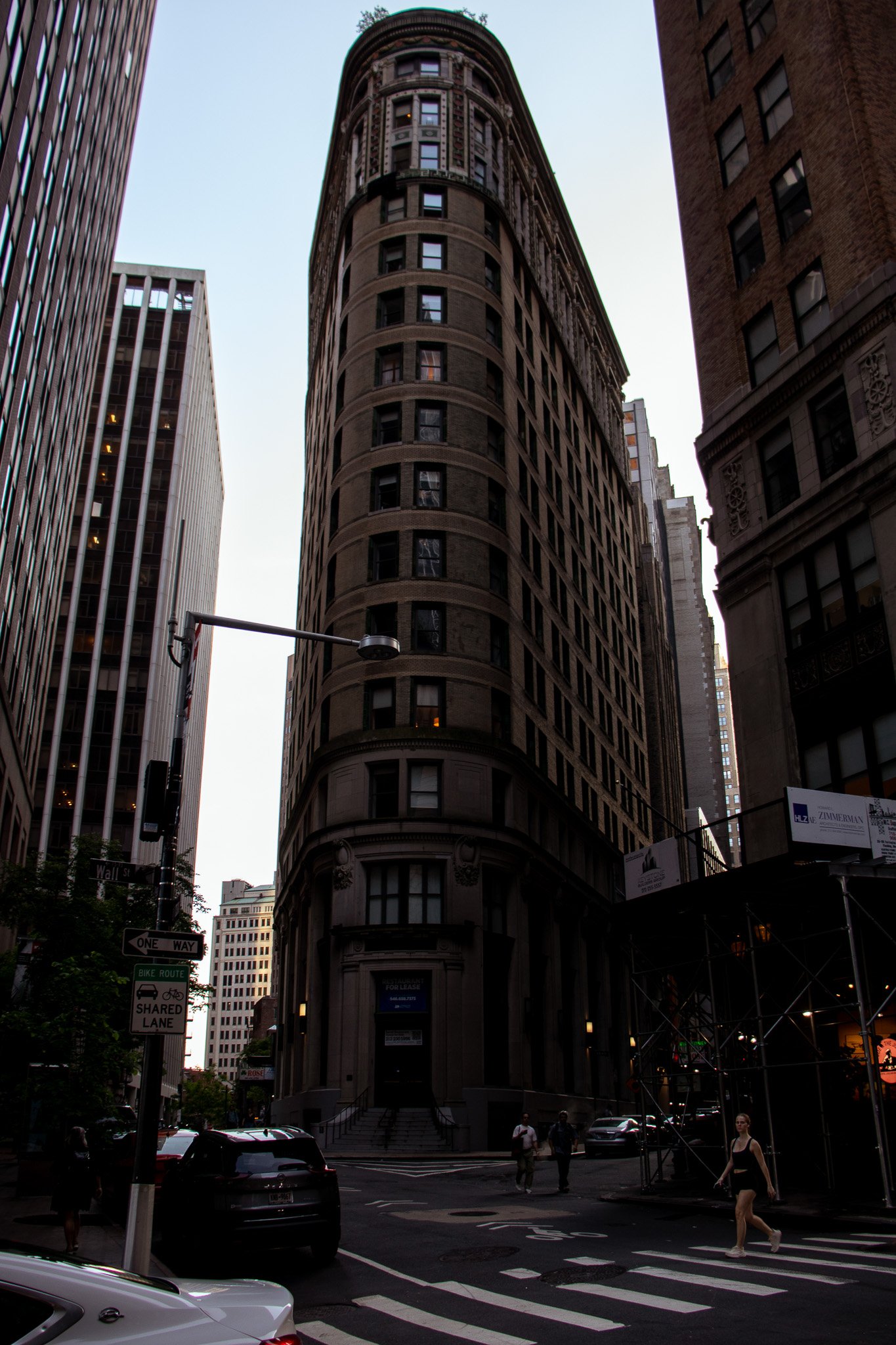 A tall, historic, rounded corner building with multiple floors and ornate architectural details, situated between modern high-rise buildings in an urban setting. The scene is at dusk, with a few pedestrians crossing the street and cars parked along t