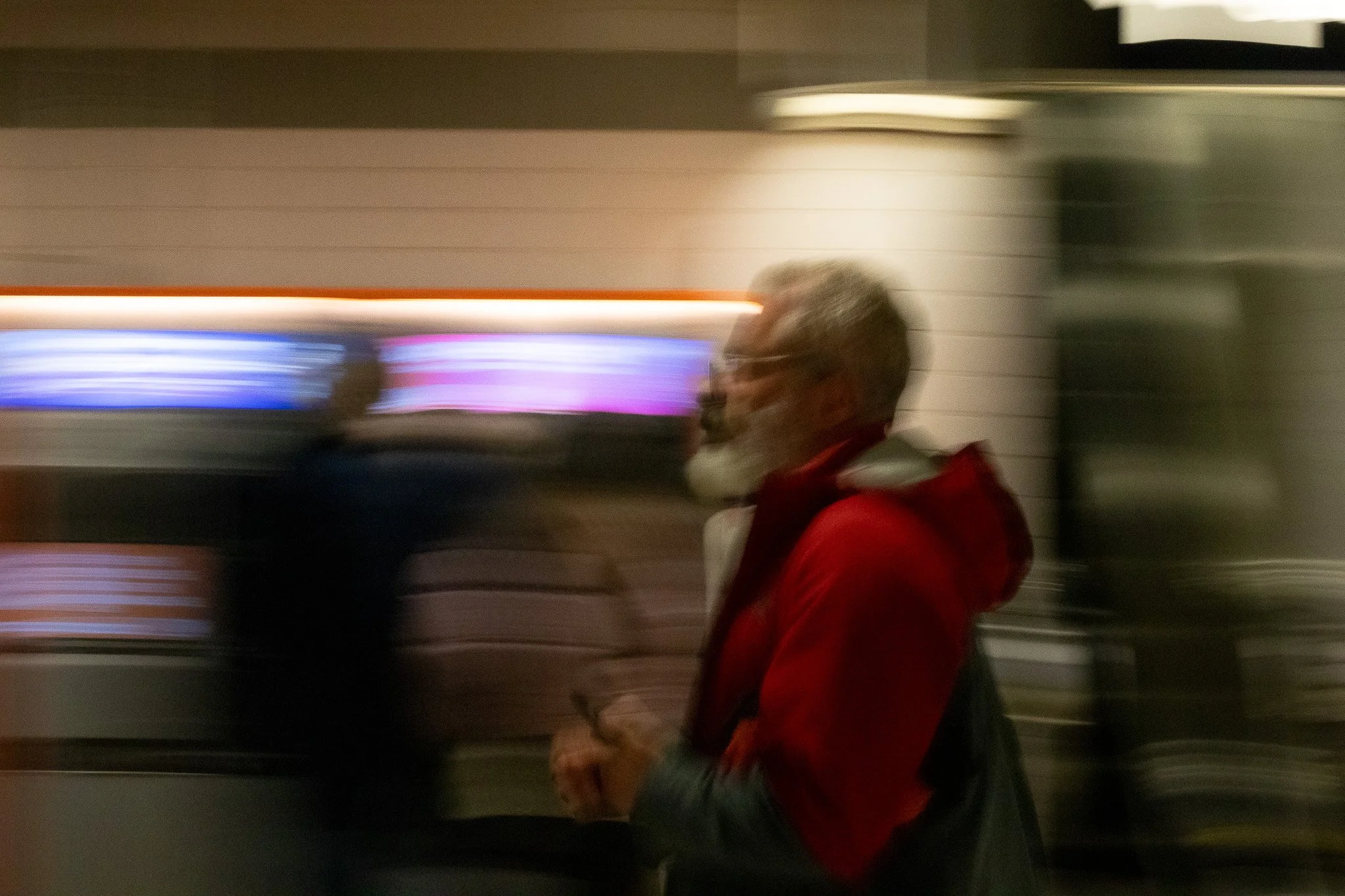 Blurry side profile of a man with gray hair, wearing glasses, a red jacket, and carrying a backpack, walking past a wall with horizontal stripes in an indoor setting.