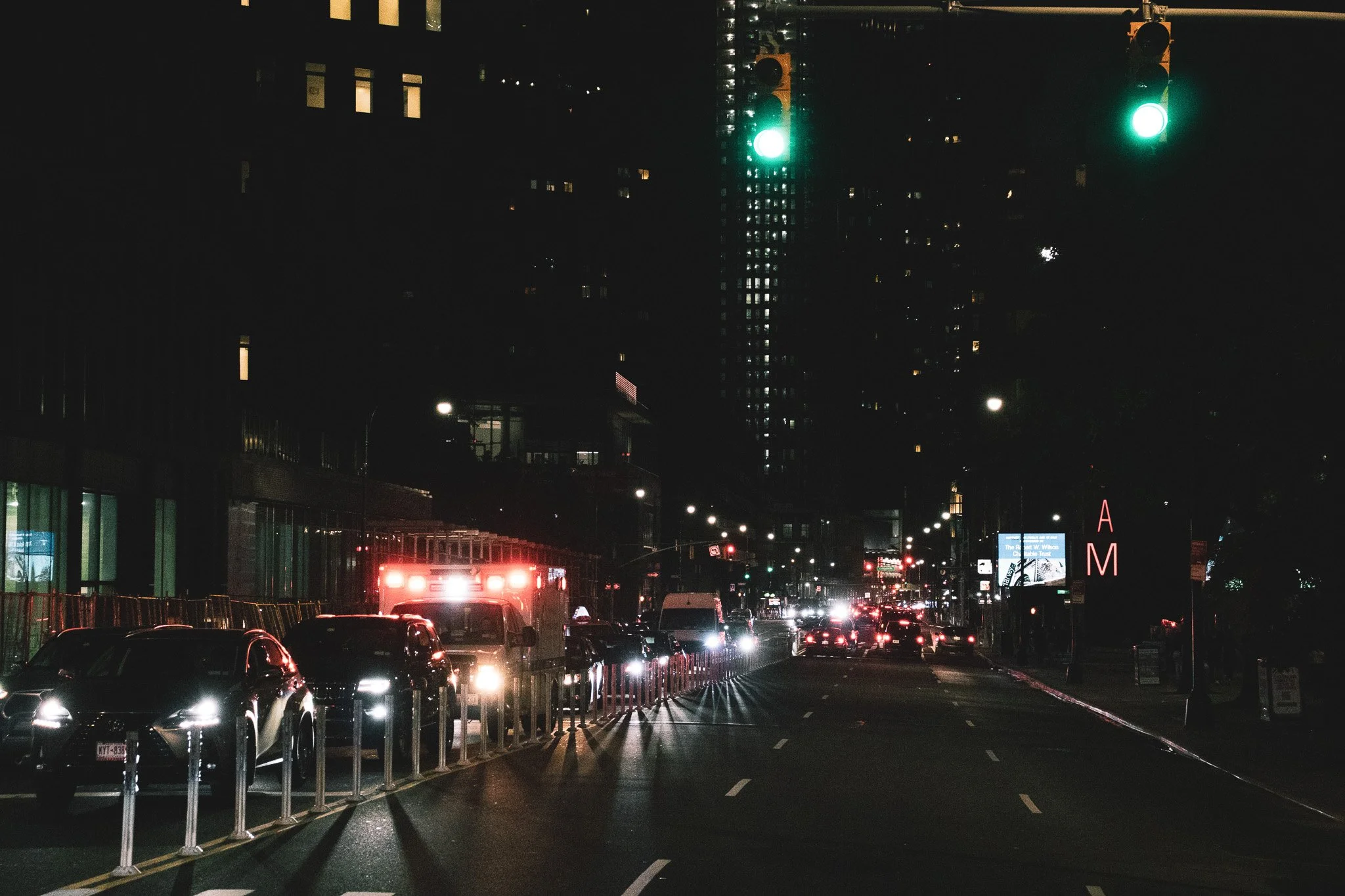 Nighttime city street with traffic, illuminated traffic lights, parked cars, and buildings with lit windows.