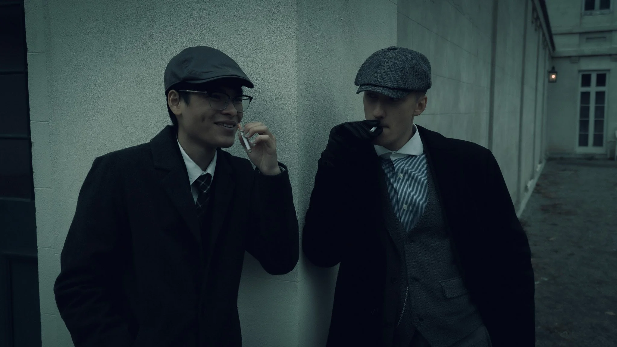 Two men in vintage-style clothing, one smiling and holding a cigarette, the other appearing serious, standing against a building wall at night.