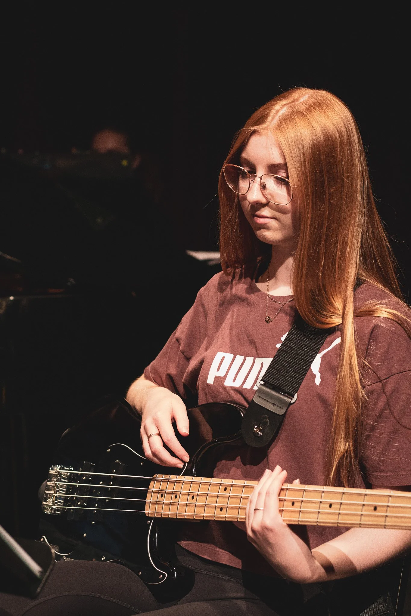 A young woman with long red hair and glasses playing an acoustic guitar, wearing a Puma shirt, in a dark room with a black background.
