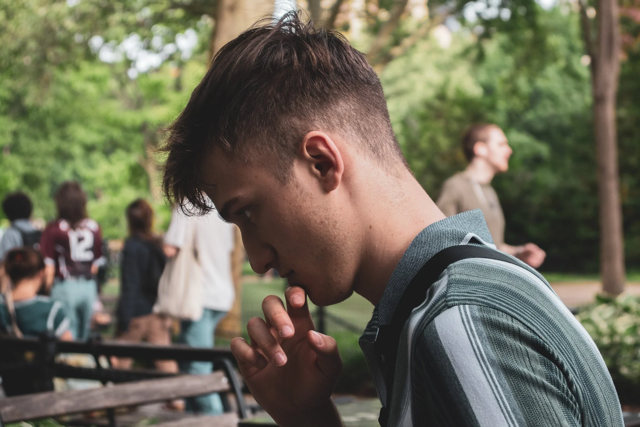 A young man with short dark hair, wearing a striped shirt, is praying outdoors with his eyes closed and hands clasped together. In the background, there are several people gathered in a park with green trees.