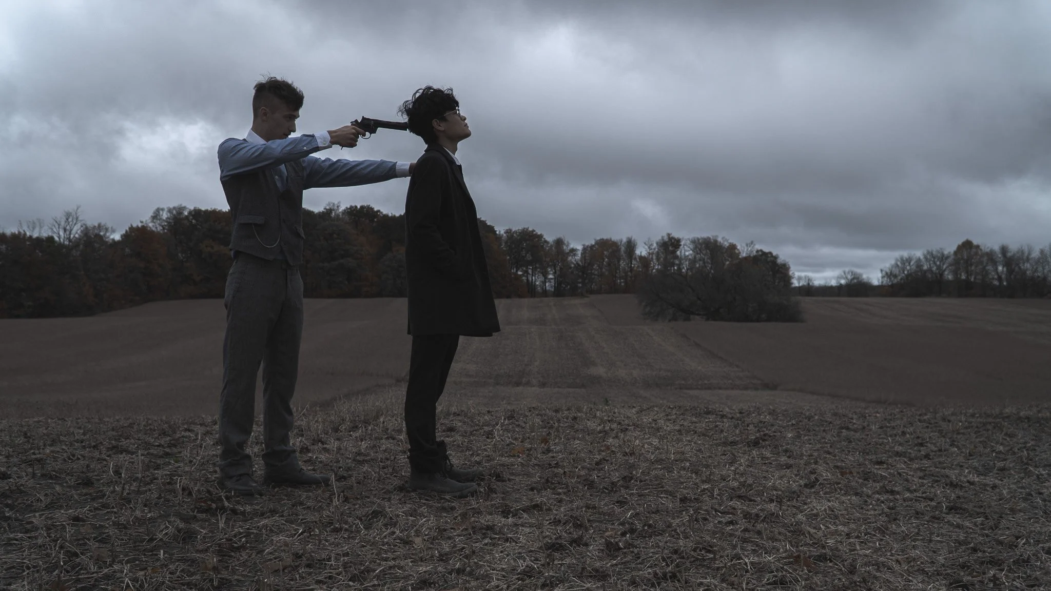 A person standing outdoors in a field on a cloudy day, with another person behind them pointing a gun at their head. The scene is dark and ominous.
