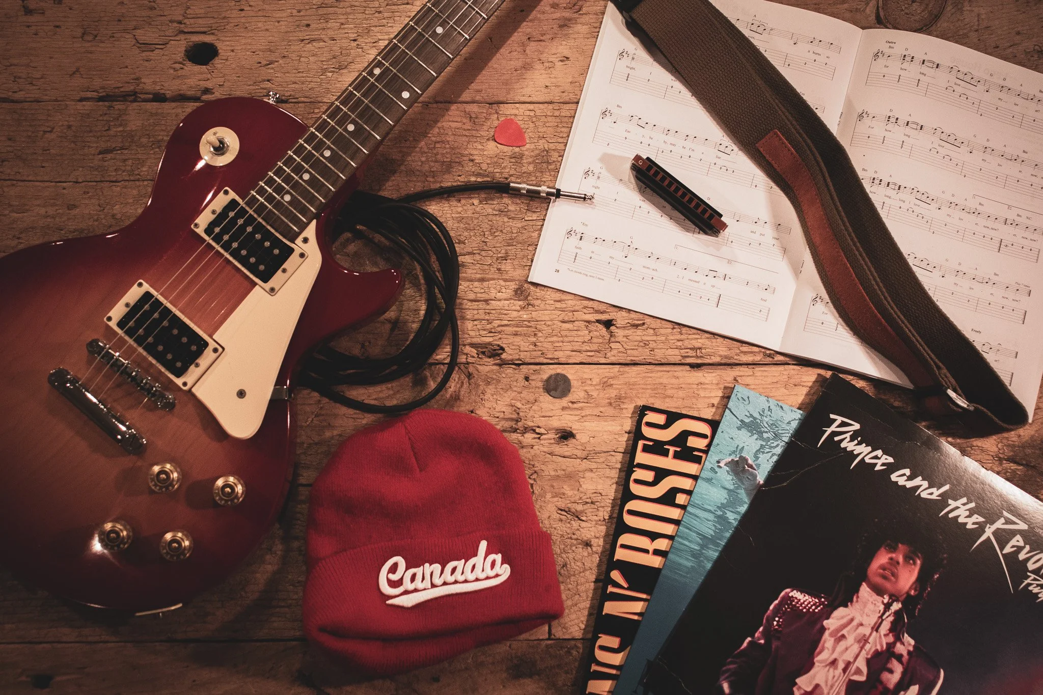 An overhead view of a rustic wooden surface with a red electric guitar, a black cable, a red Canada baseball cap, sheet music, a harmonica in a case, a magazine, and a book.