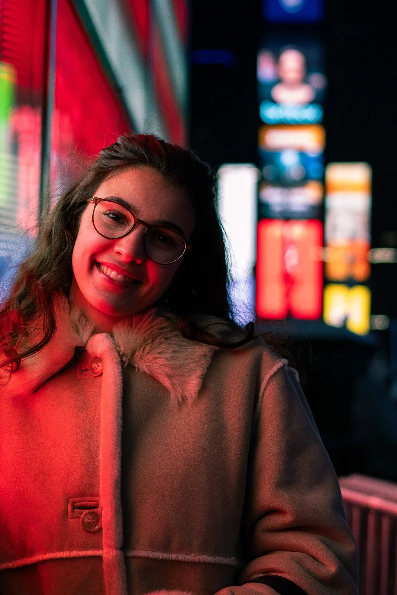 A woman with glasses smiling at night in front of brightly lit illuminated signs, wearing a tan coat with a fur collar.