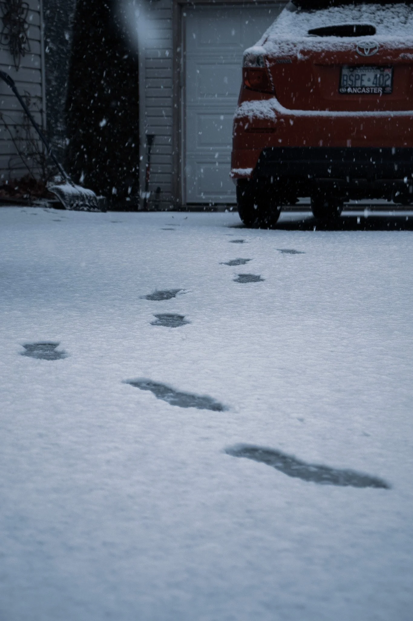 Footprints in freshly fallen snow leading to a parked red car with snow on its surface, located next to a garage and house exterior during snowfall.