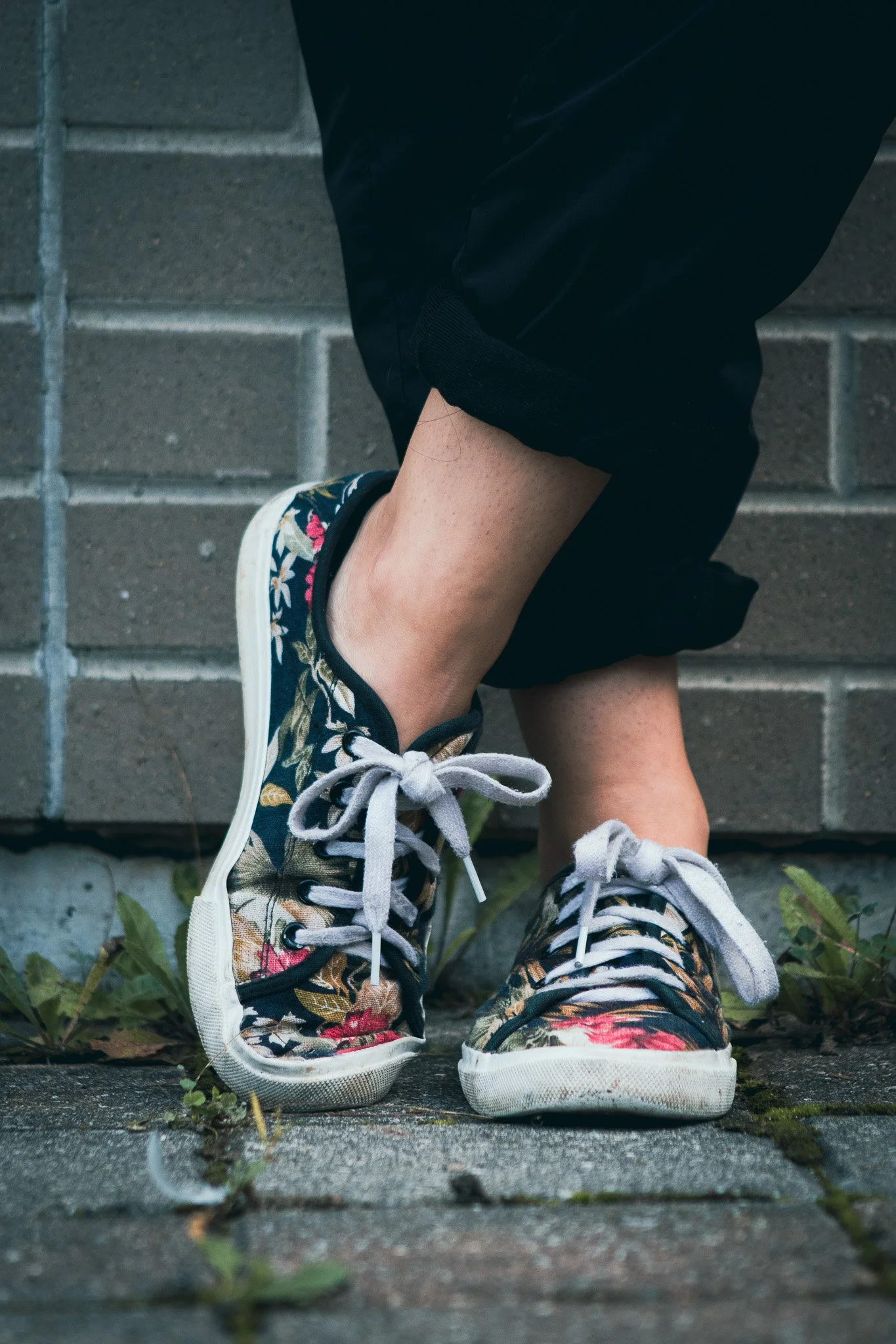 Close-up of a person's legs crossing at the ankles, wearing floral-patterned sneakers with white soles, black trim, and gray laces, standing on a brick sidewalk with small green weeds growing between the bricks, against a gray brick wall background.