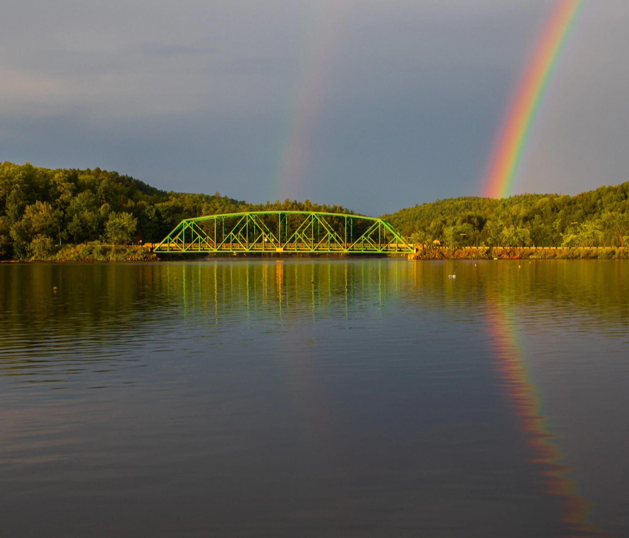 A scenic view of a river with a green bridge crossing over it, surrounded by lush green hills and a colorful rainbow in the cloudy sky.
