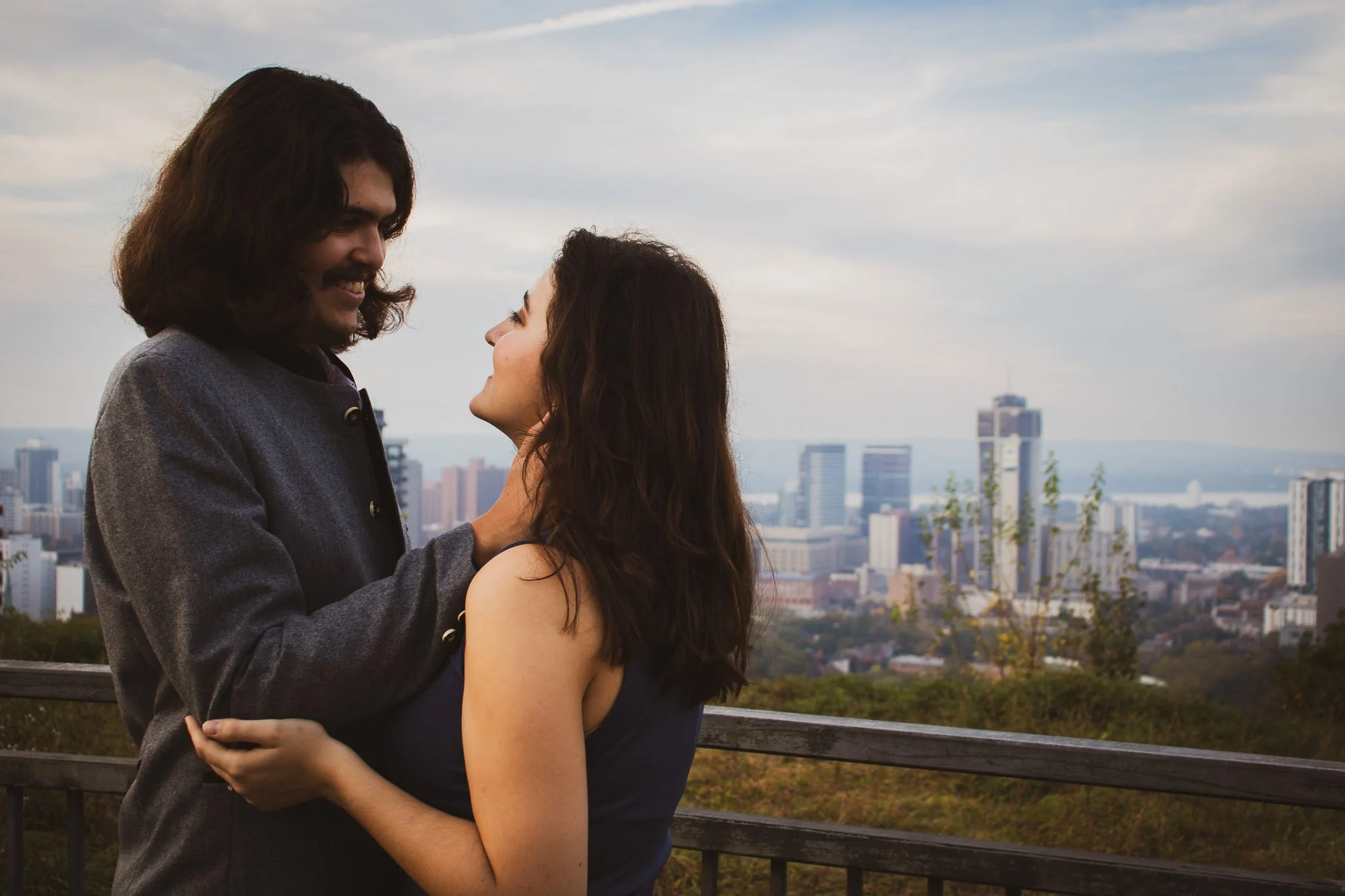 A man and woman are standing close together on a balcony with a city skyline in the background. They are looking into each other's eyes and smiling, holding each other affectionately. The man has shoulder-length dark hair and a mustache, wearing a gr