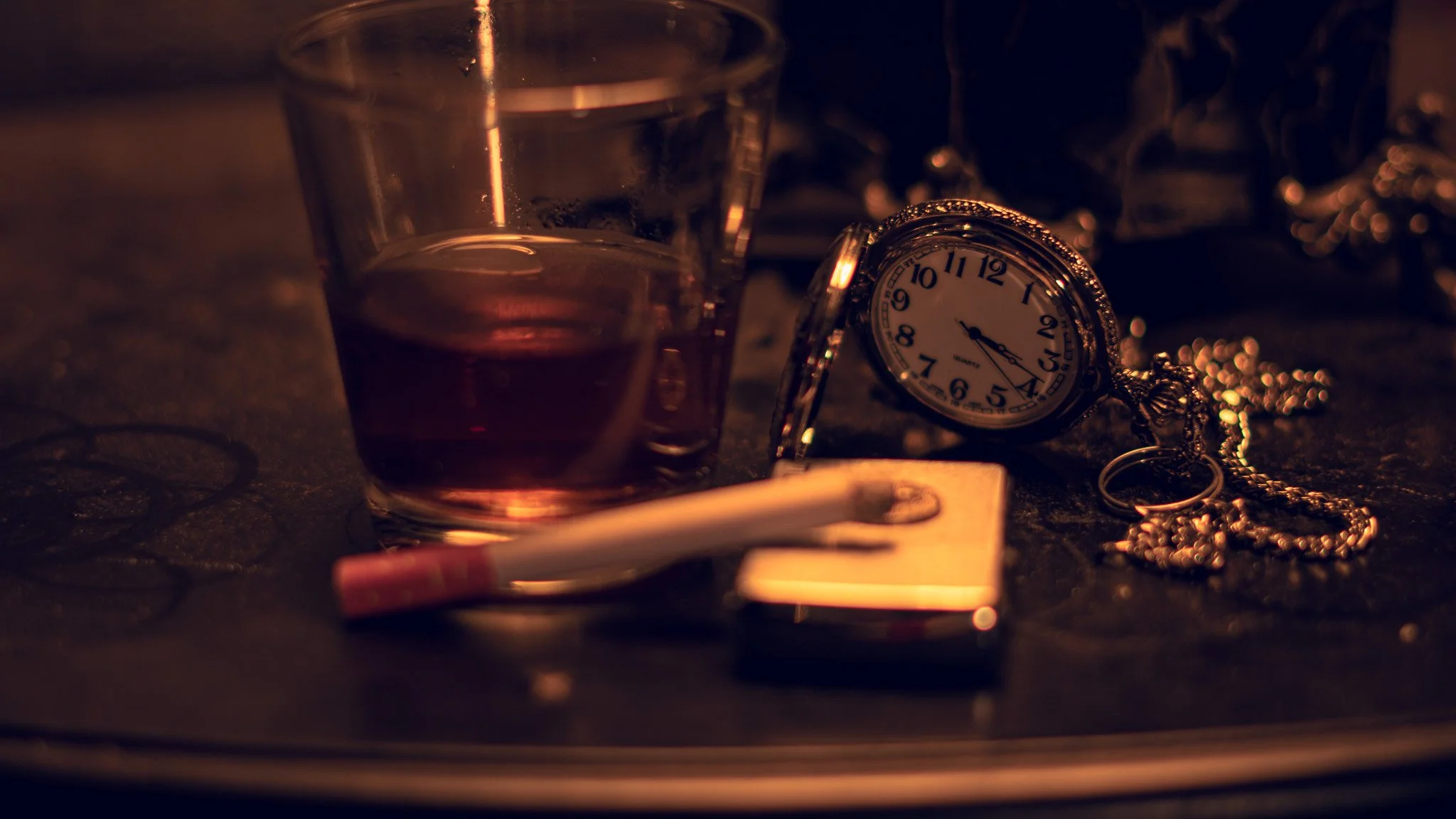A close-up of a glass of alcohol, an antique pocket watch, and scattered jewelry on a dark surface, with warm, dim lighting.