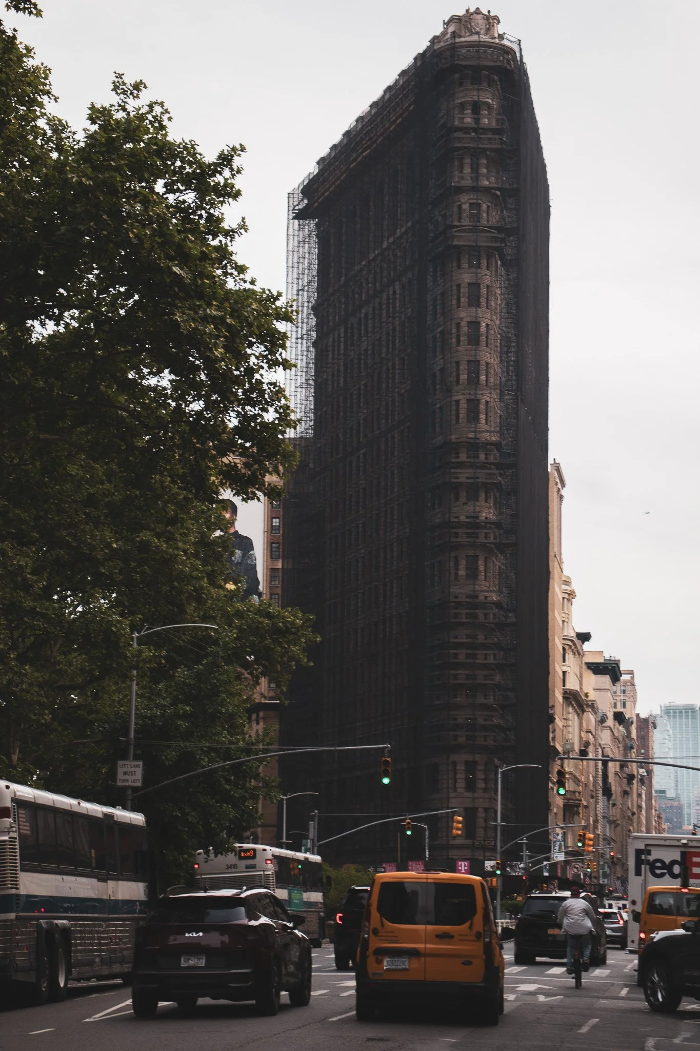 A city street scene with traffic, including buses, cars, and a yellow taxi. A tall, dark building with ornate architecture and scaffolding is in the background, and trees line the street.