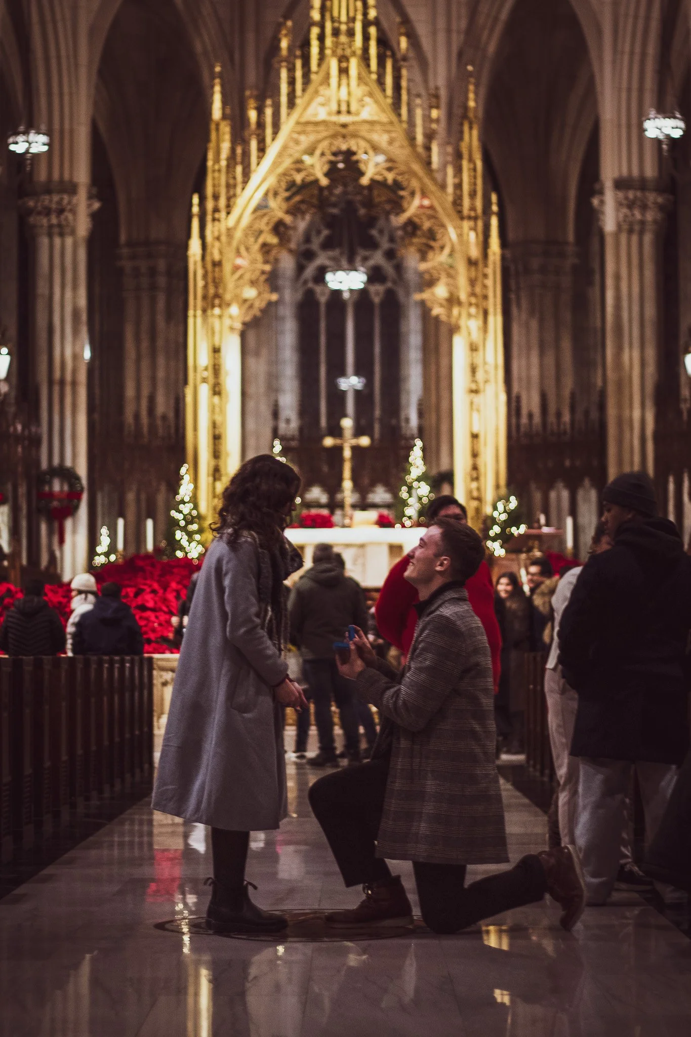 A man proposing marriage to a woman inside a church, with the woman standing and the man kneeling on one knee. They are surrounded by other people and Christmas decorations.