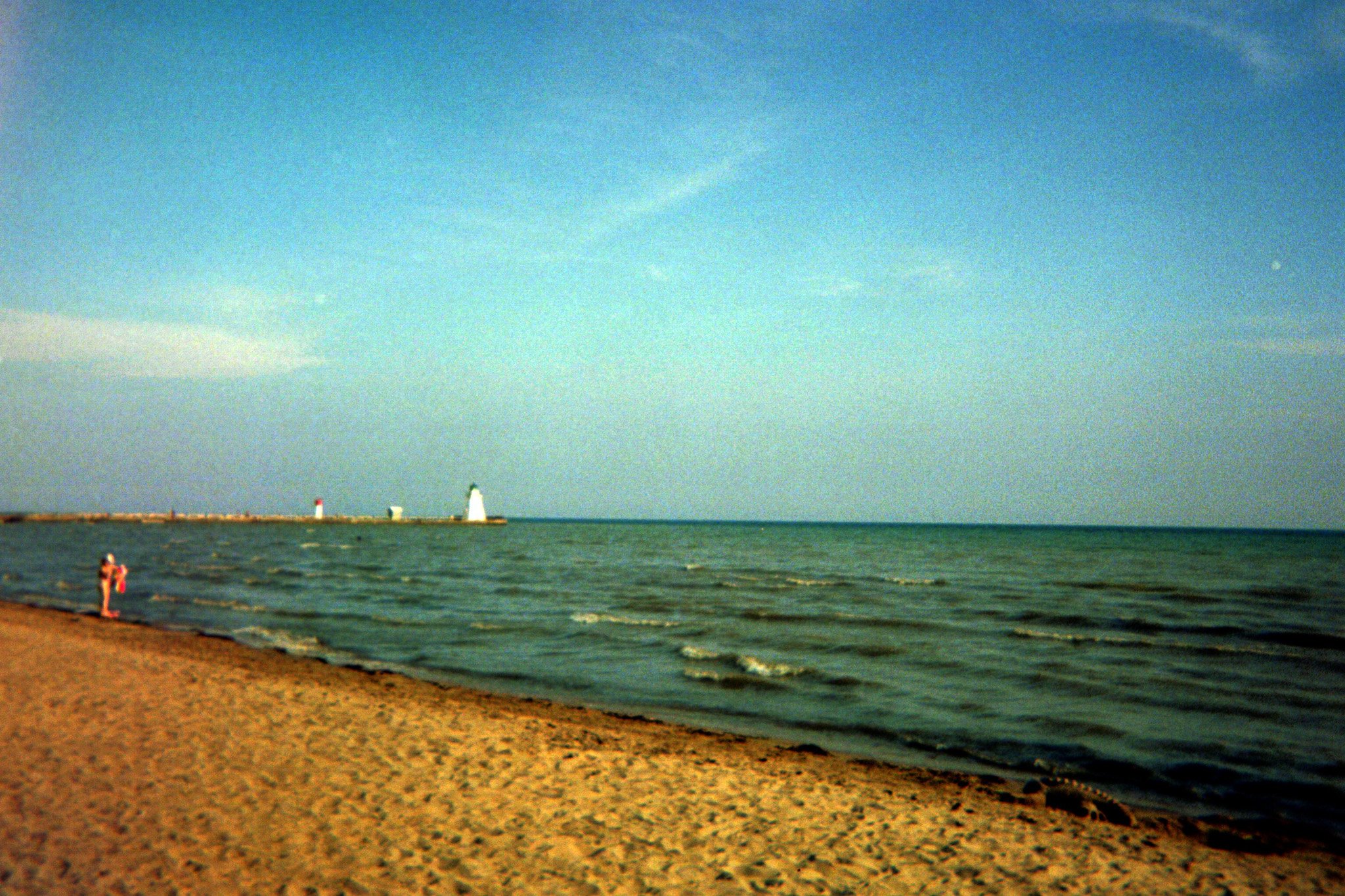 A beach scene with a sandy shoreline, two children playing near the water, and a lighthouse or navigational aid structure on a breakwater in the distance. The sky is partly cloudy with a small bright moon visible.