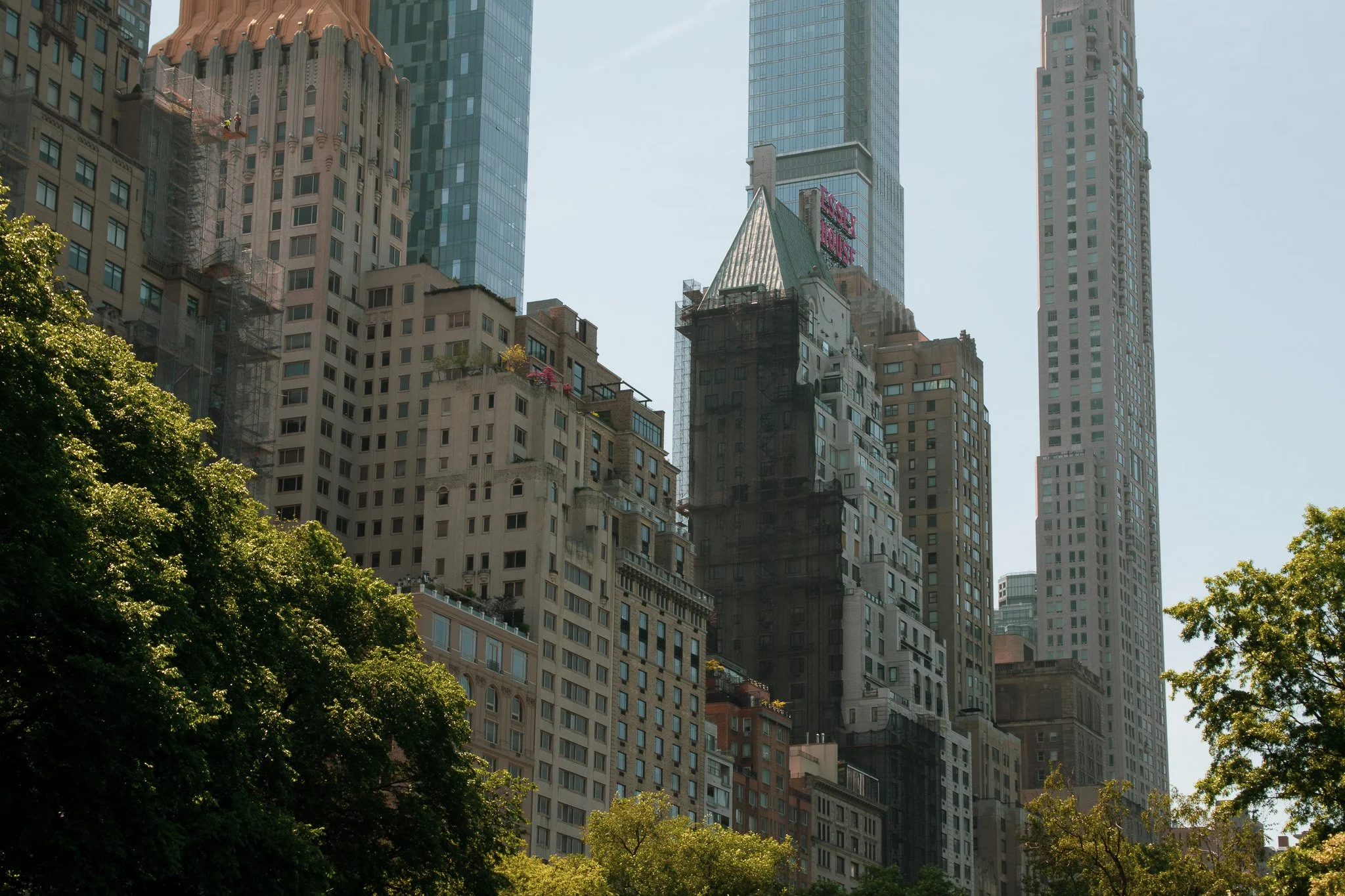 City skyscrapers with a mix of modern glass buildings and older architecture, with green trees in the foreground and a clear blue sky.