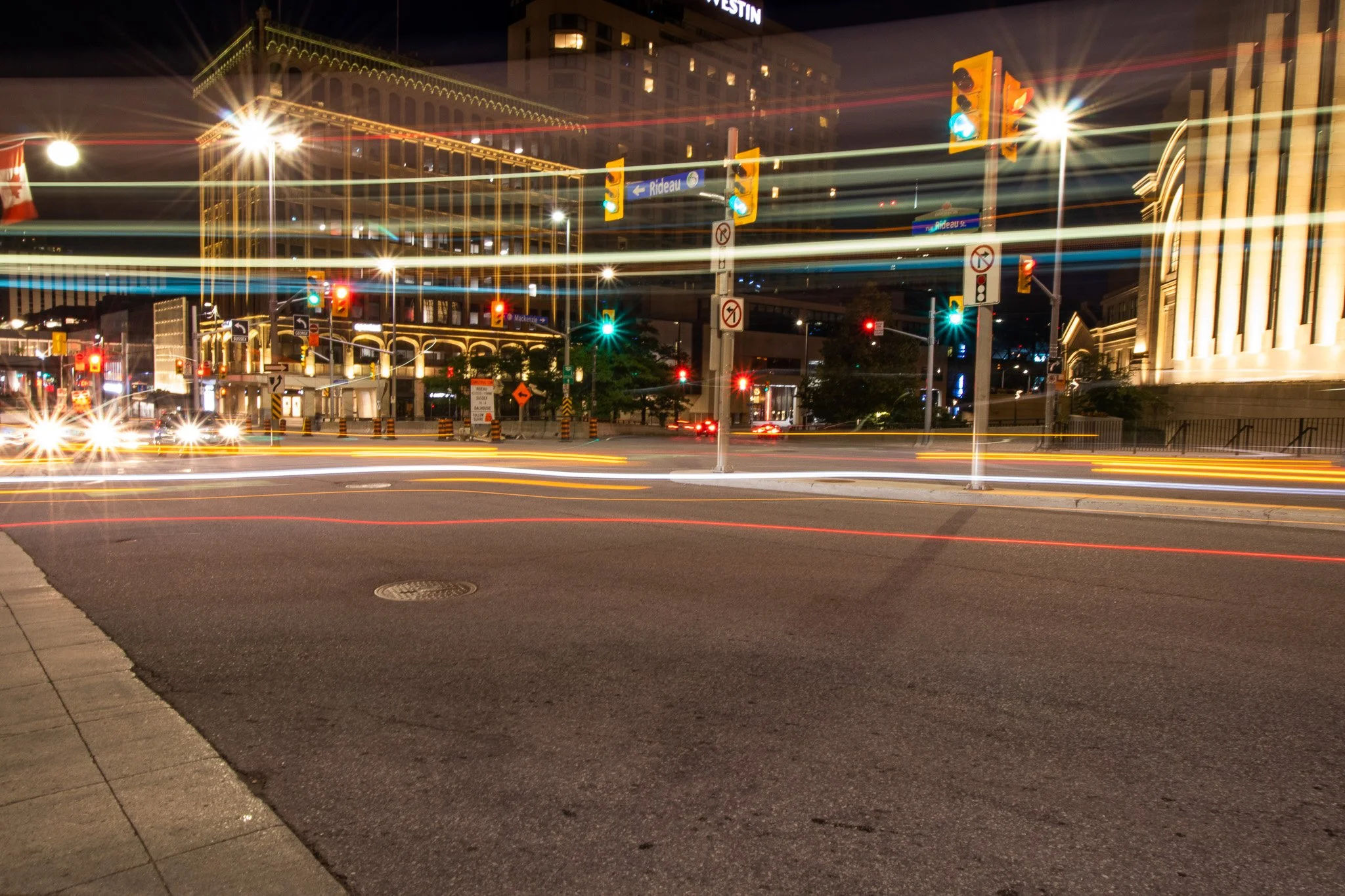 Nighttime city street scene with traffic lights, light trails from moving vehicles, and illuminated buildings in the background.