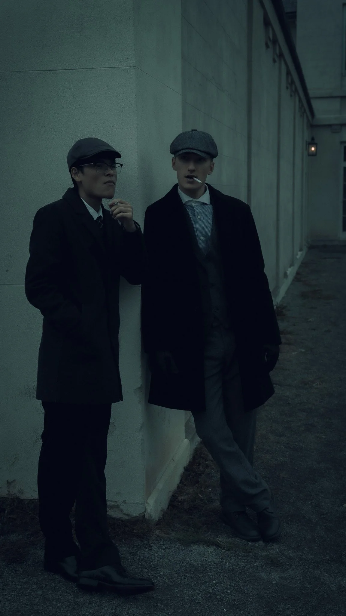 Two young men in vintage clothing standing against a wall at dusk, one smoking a cigarette and wearing a flat cap, the other with glasses and a newsboy cap.