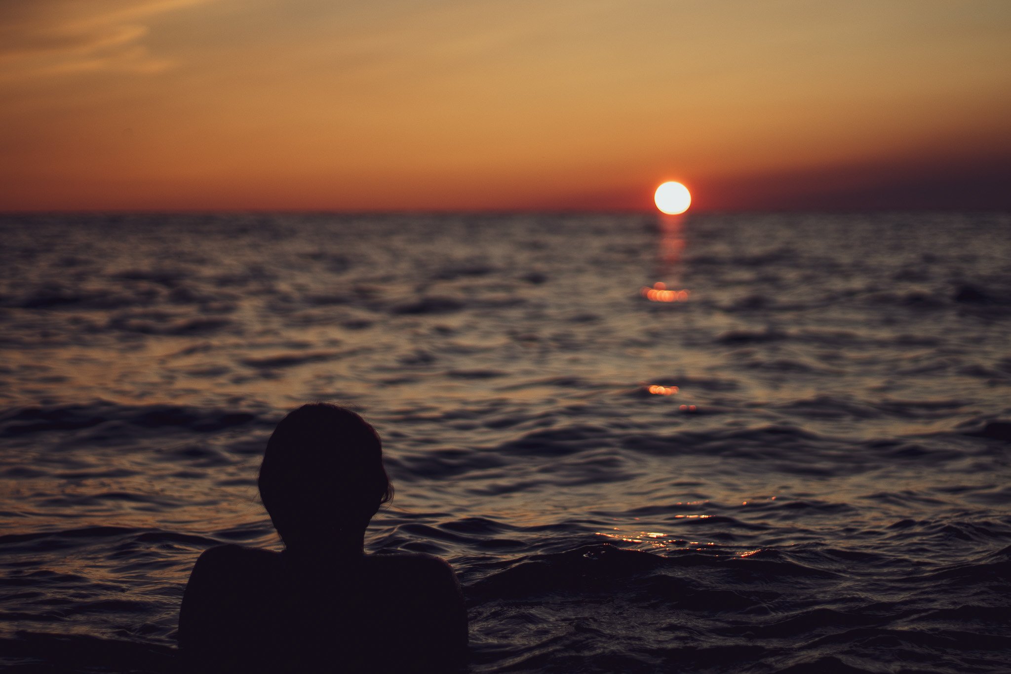 A person with long hair is swimming in the ocean during sunset, with the sun on the horizon and a colorful sky.