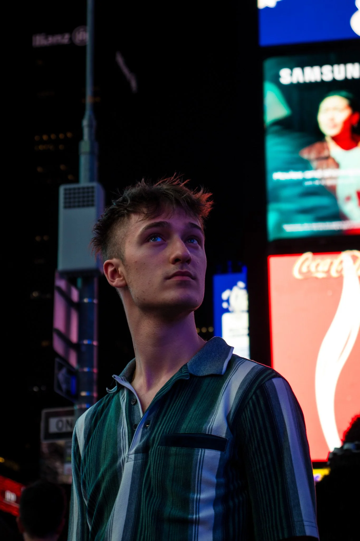 A young man looking upward in a city at night, illuminated by bright digital billboards and advertisements, including one for Samsung, with a blurred cityscape in the background.