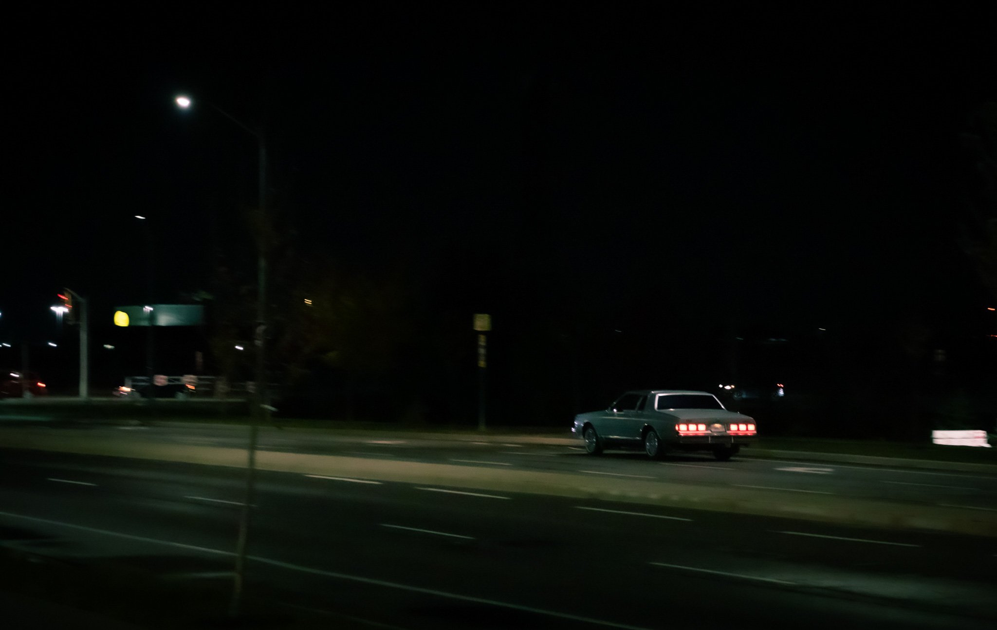 Nighttime parking lot scene with a silver car, some parked vehicles, and illuminated streetlights in the background.