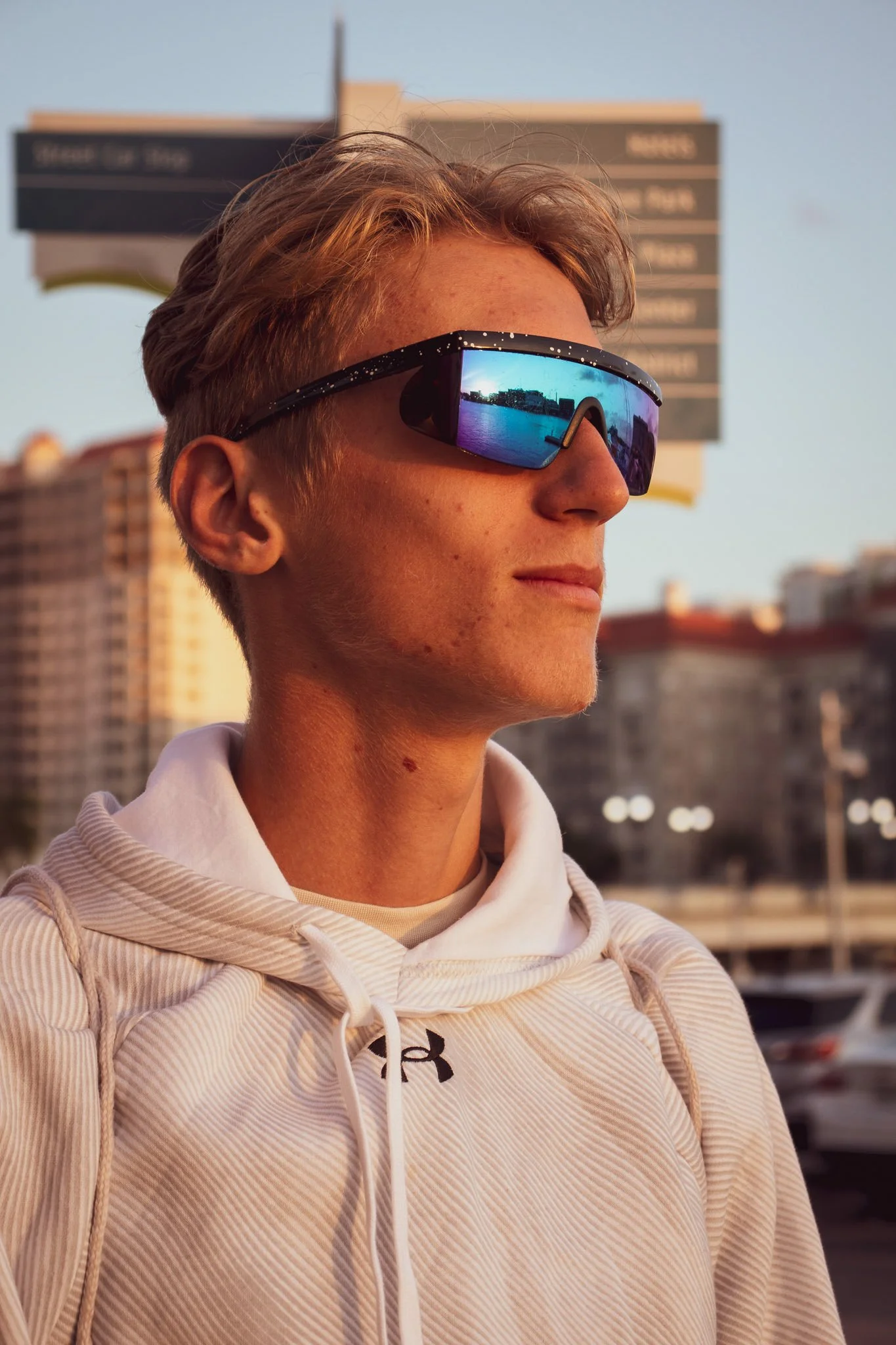 Young man in a white hoodie wearing reflective sunglasses over a city skyline during sunset.