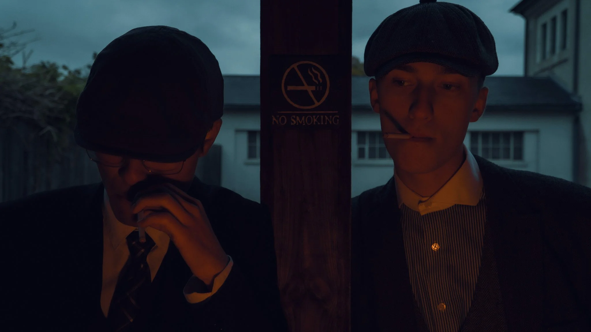 Two young men dressed in vintage clothing, one smoking, standing behind a no smoking sign on a wooden post during dusk.