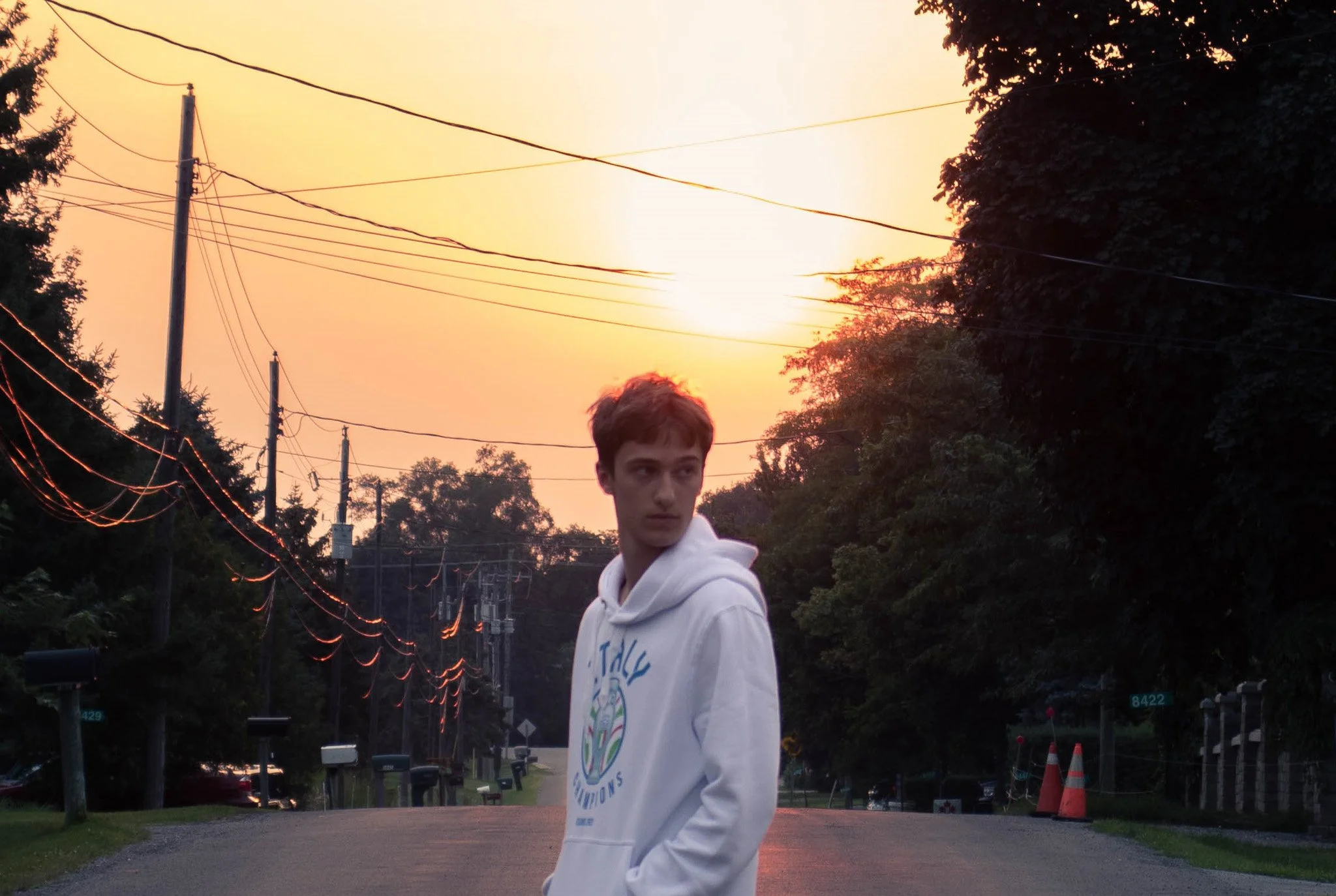 A young man in a white hoodie standing outdoors during sunset with power lines, trees, and suburban houses in the background.