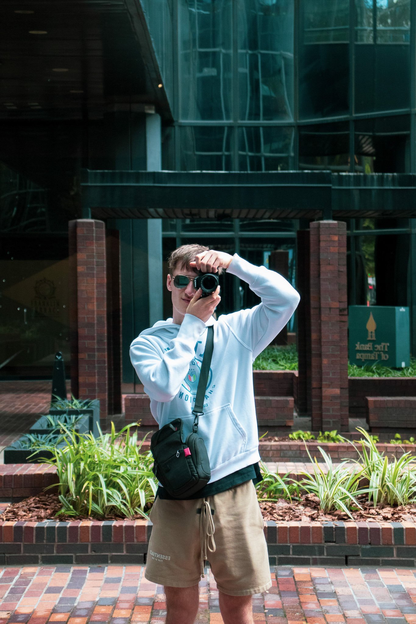 Young man taking a selfie in front of a modern glass building, wearing sunglasses, a white hoodie, and beige shorts, with a crossbody bag, standing near plants and brick pavement.