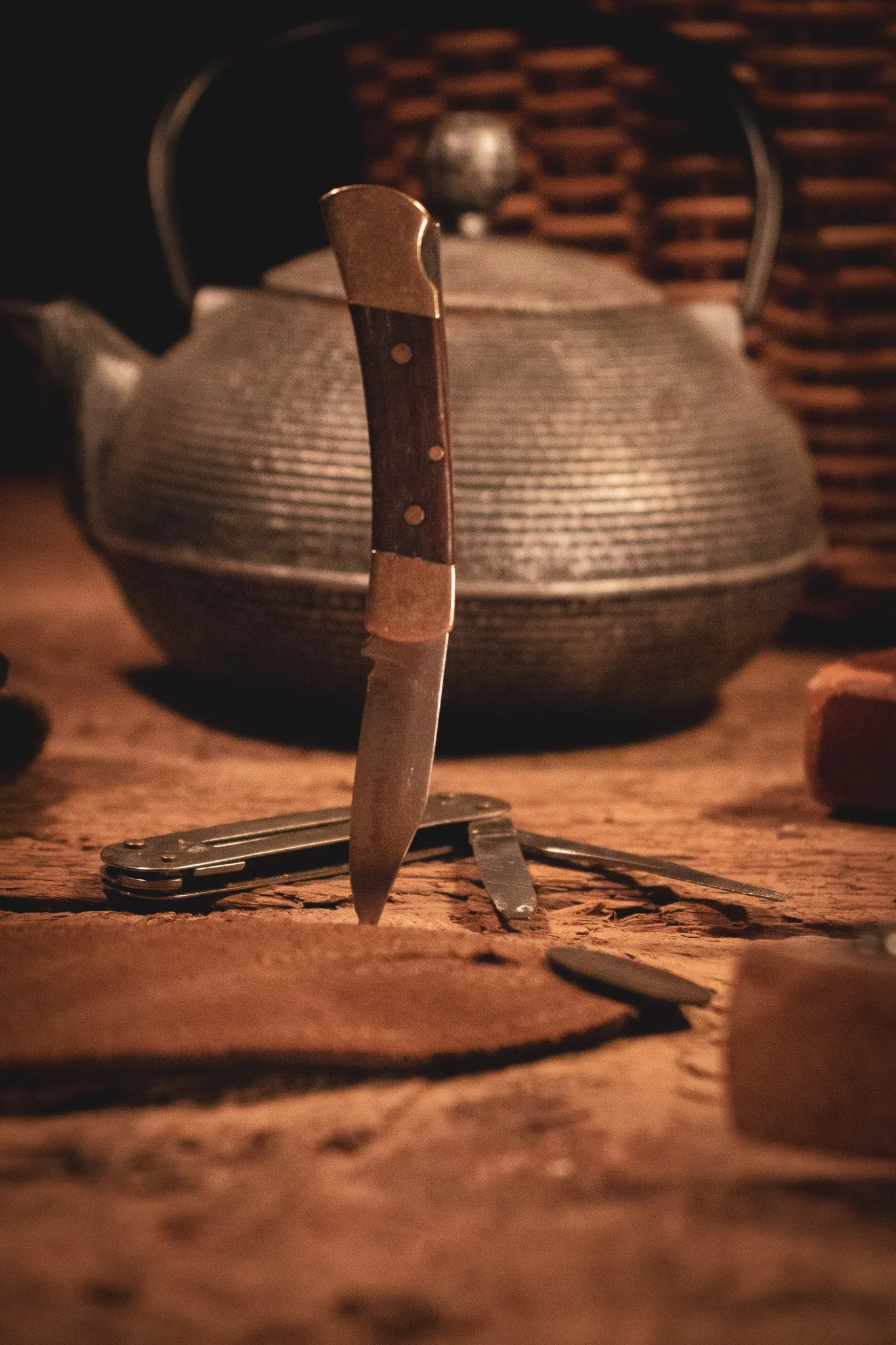 A pocket knife is standing upright on a rough wooden surface with a folding knife open in front of it. In the background, there is a traditional metal teapot with a woven basket behind it.