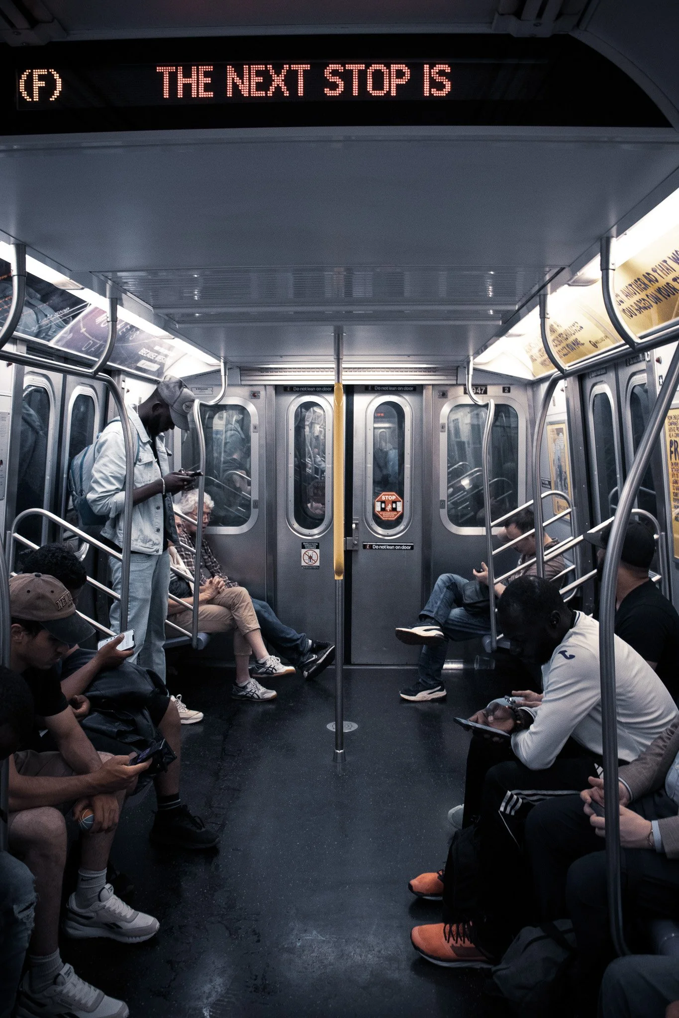 Inside a subway train with passengers seated and standing, some using their phones, with a digital sign above reading 'The next stop is'.