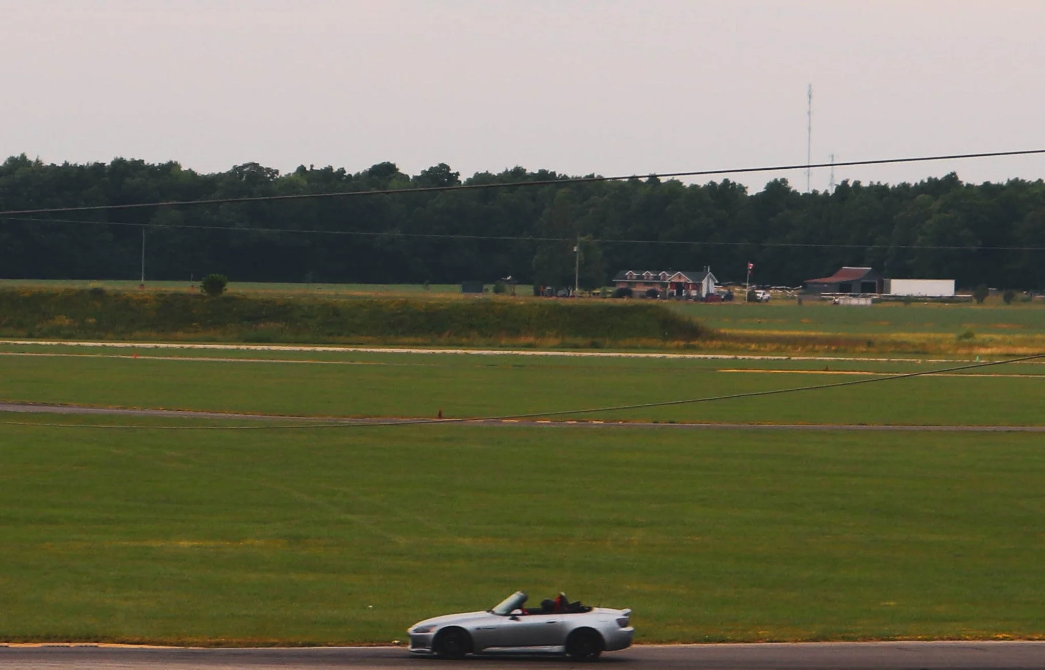 A white convertible sports car driving on a rural road with grassy fields, trees, and farm buildings in the background.