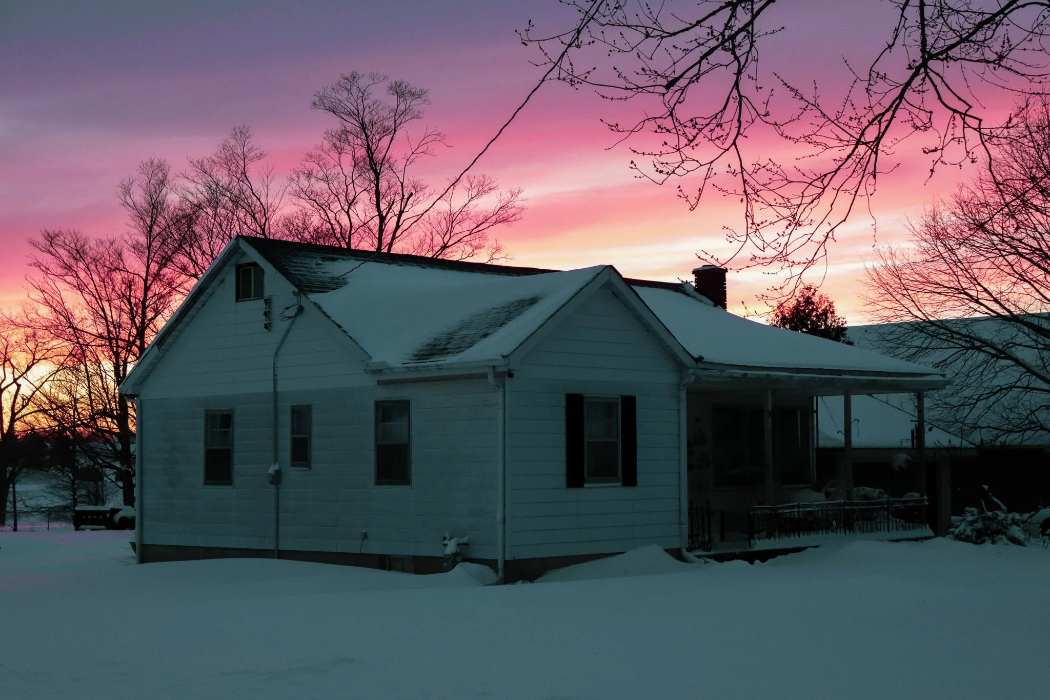 Snow-covered backyard and house against a colorful sunset sky with bare trees.