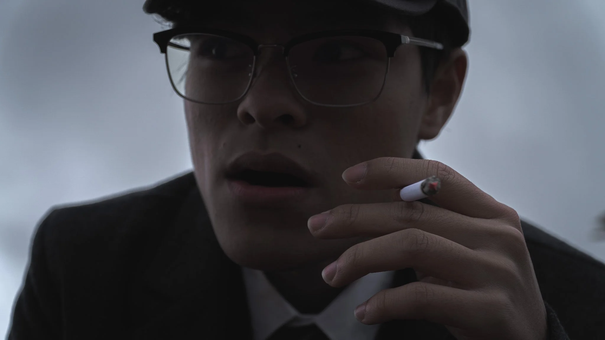 A young man wearing glasses holding a cigarette close to his mouth, with a serious expression, in an outdoor setting with a cloudy sky.