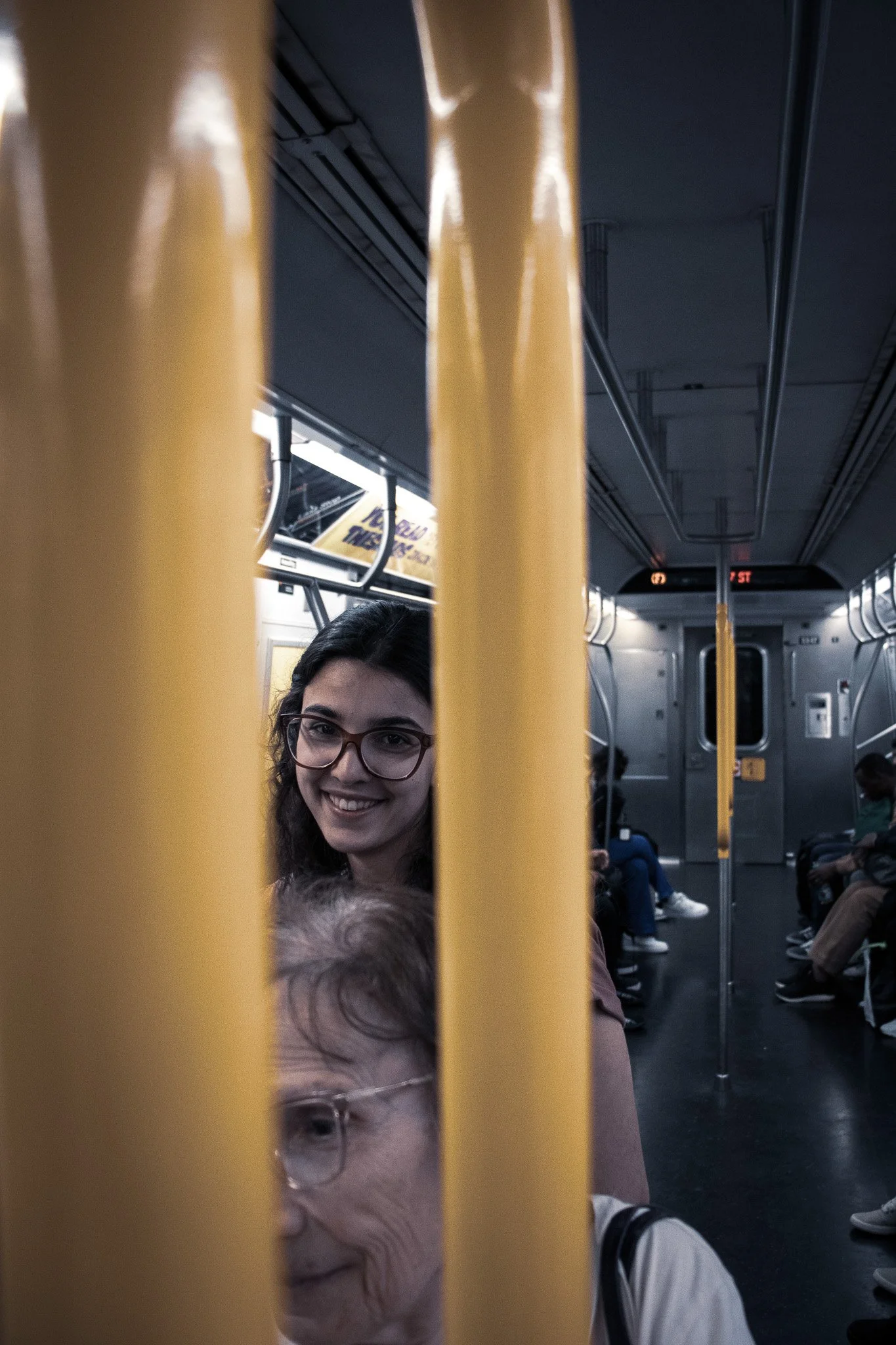 People sitting inside a subway train, seen through yellow poles dividing the view, with some faces visible and others obscured, including a woman with glasses smiling.