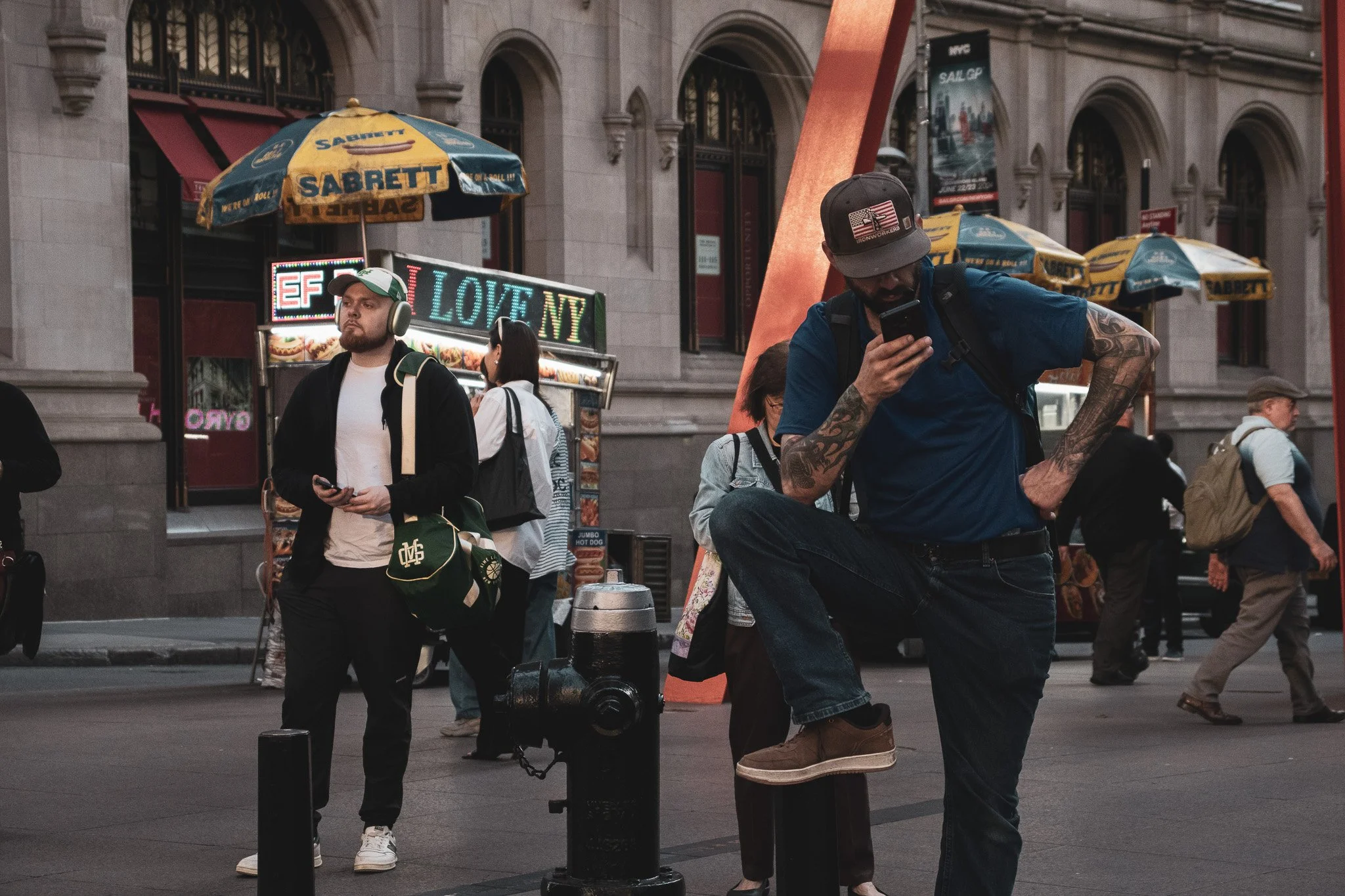 People walking and standing on a city street with historic building facades, colorful umbrellas, and neon signs, including one that reads 'LOVE NY'.