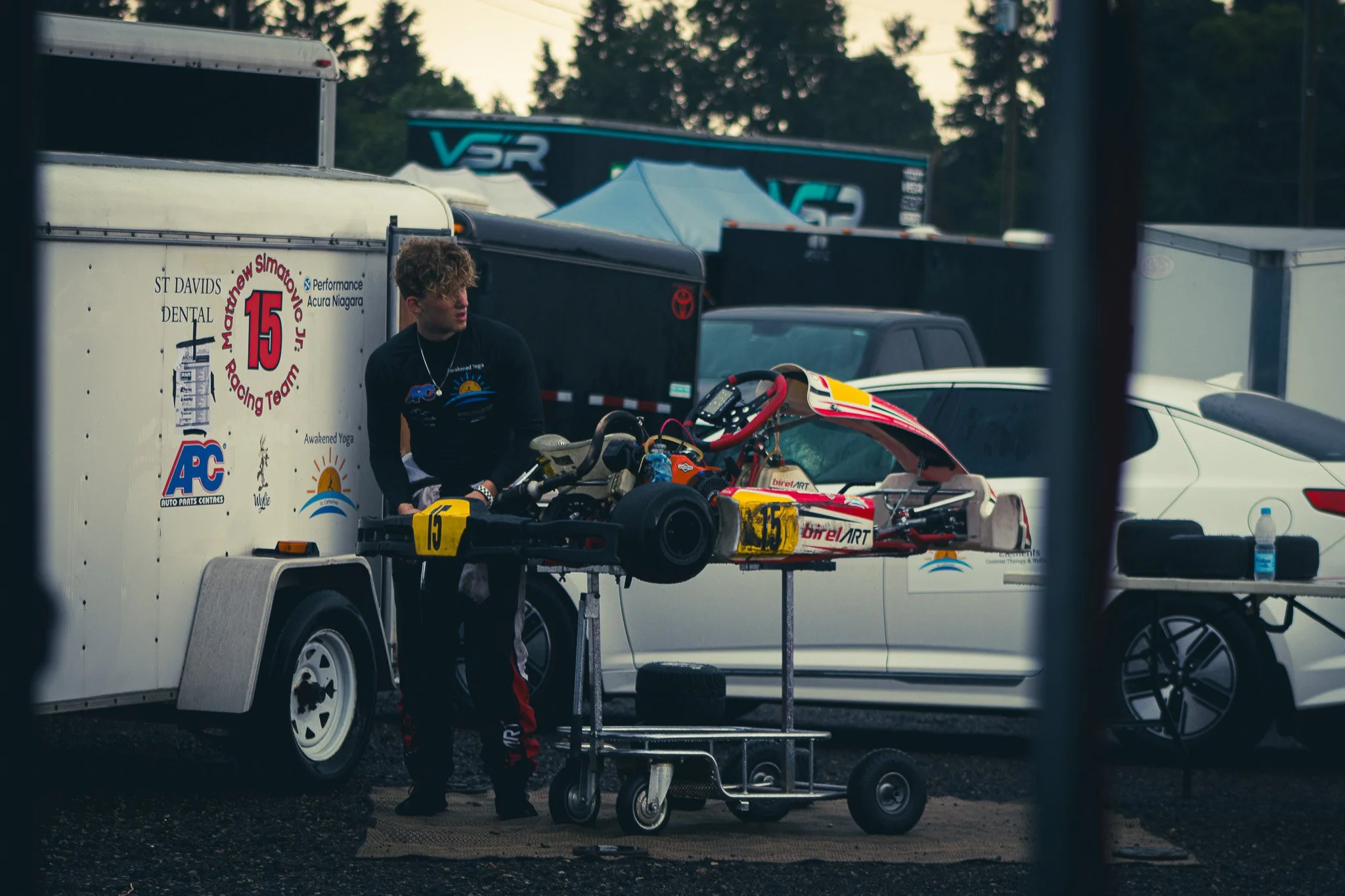 A young man with curly hair and black and red racing suit standing next to a racing go-kart on a cart in a parking lot, with trailers and cars in the background.